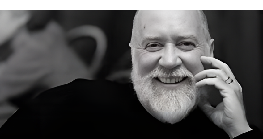 A smiling man with a white beard, bald head, wearing a dark shirt, resting his hand on his face with a ring on his finger.