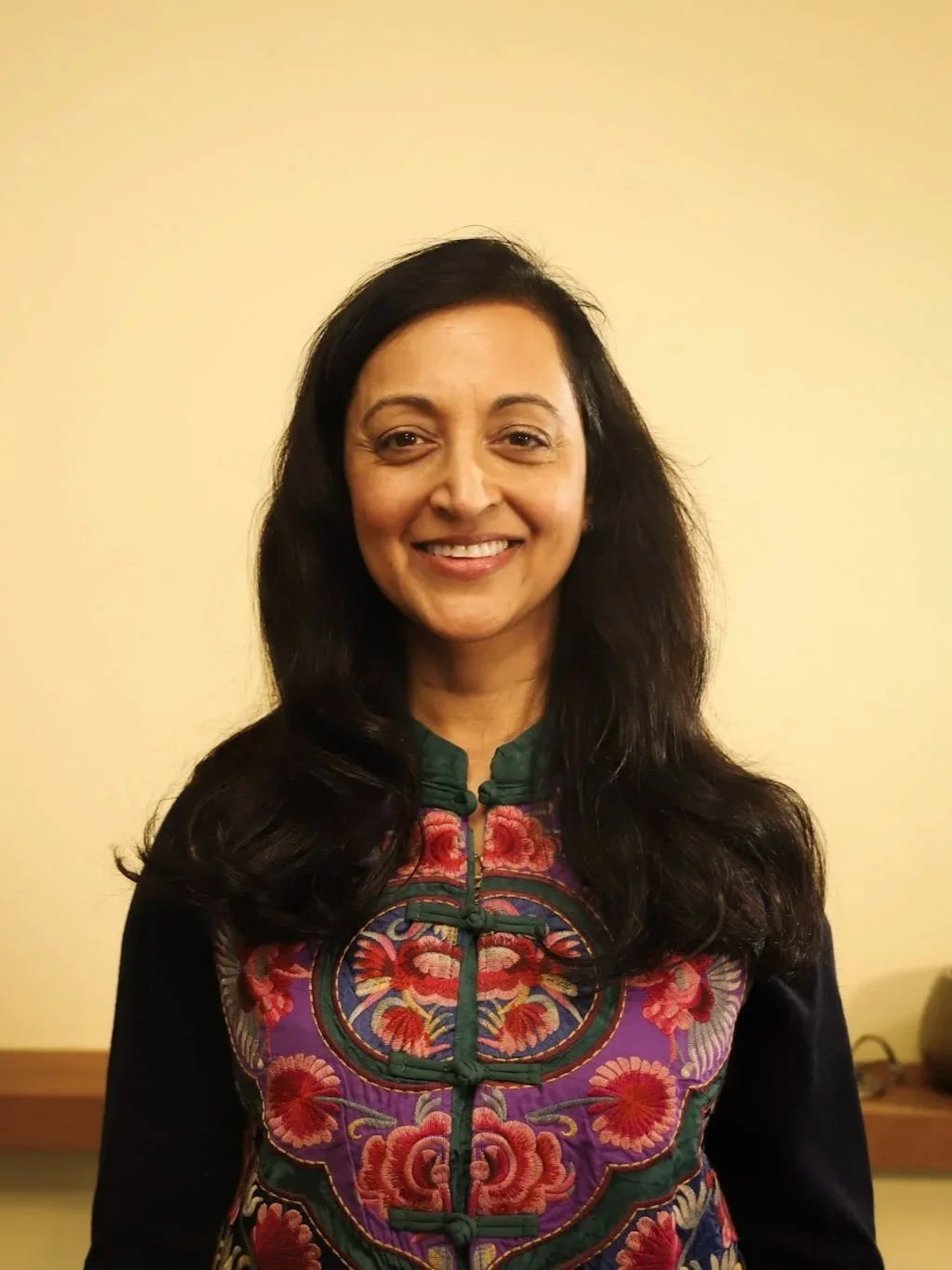 A woman with long dark hair smiling, wearing a colorful embroidered top with floral patterns, standing against a beige wall.