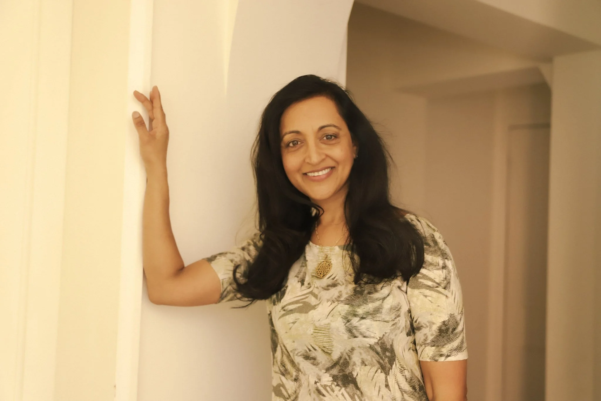 A woman with long dark hair smiling, standing in a hallway with her hand touching a white wall, wearing a patterned top and a pineapple-shaped necklace.
