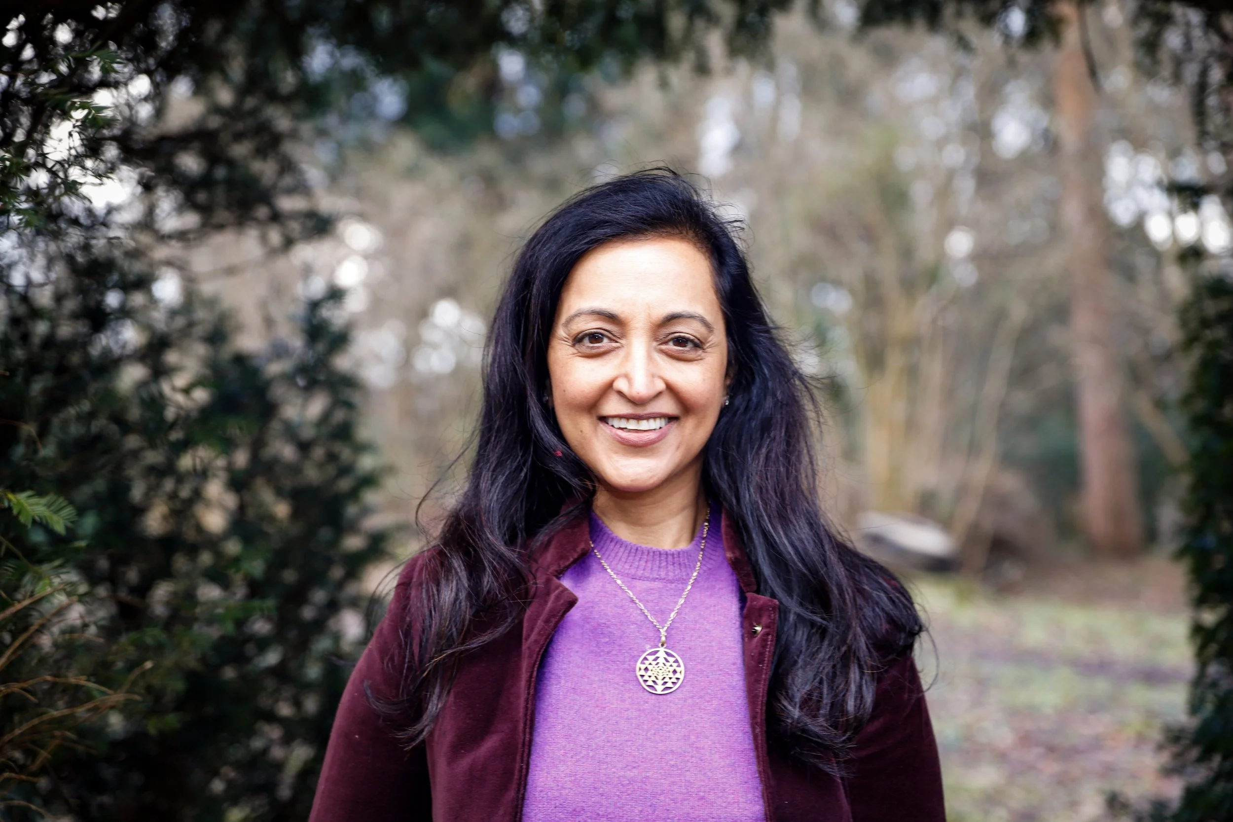Roxana with long dark hair smiling outdoors in a wooded area, wearing a purple sweater, a maroon jacket, and a gold pendant necklace.
