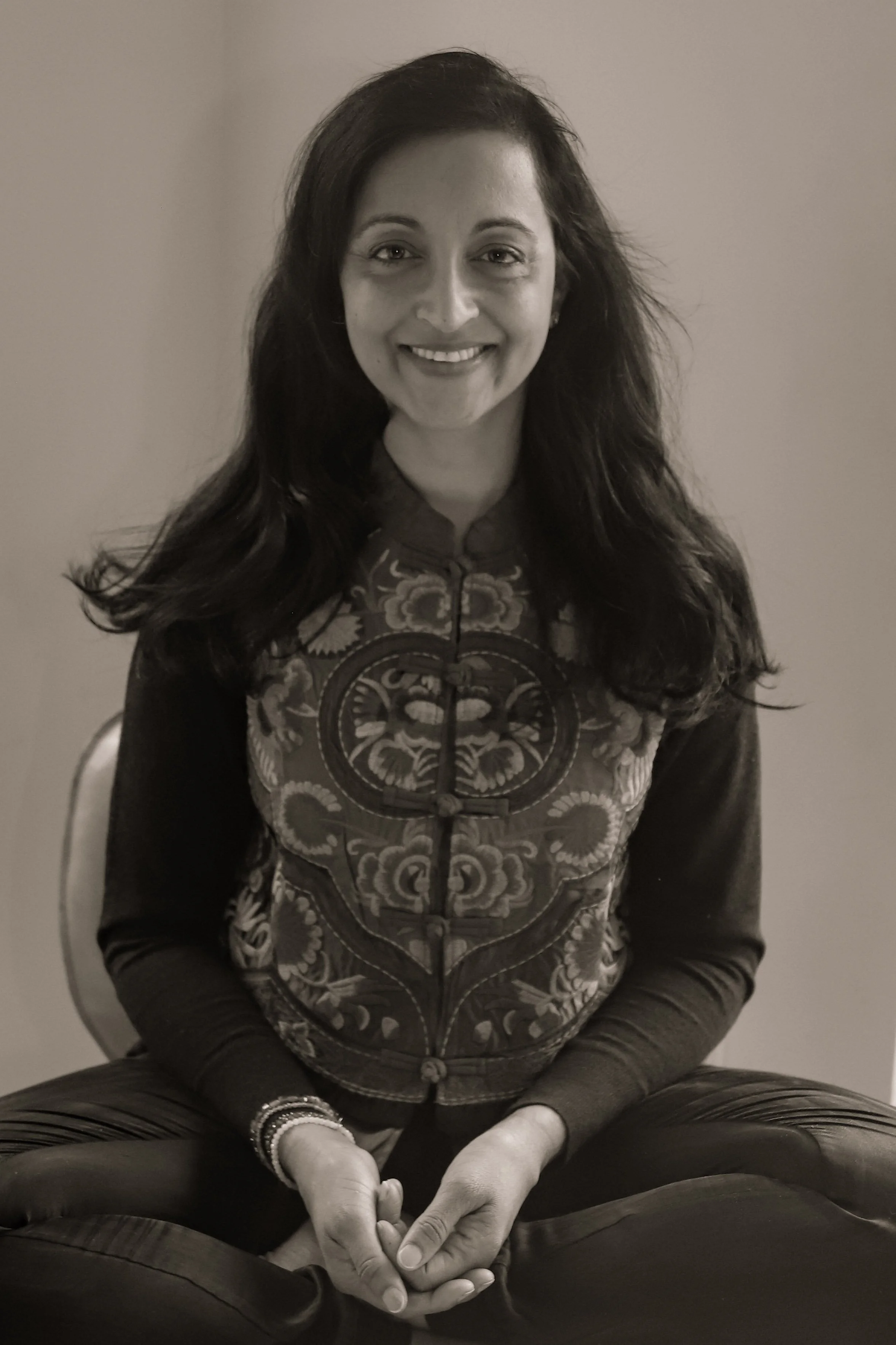 A woman with long dark hair smiling, seated with legs crossed, wearing a patterned jacket and bracelets on her left wrist.