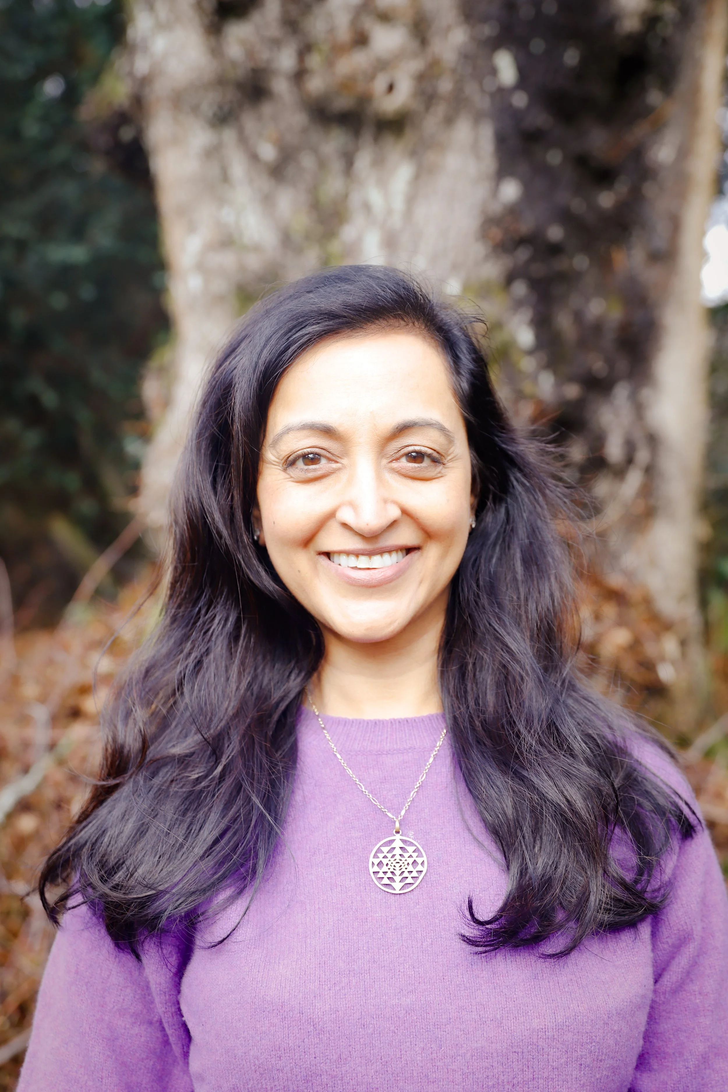 A woman with long dark hair smiling outside near a tree, wearing a purple top and a necklace.