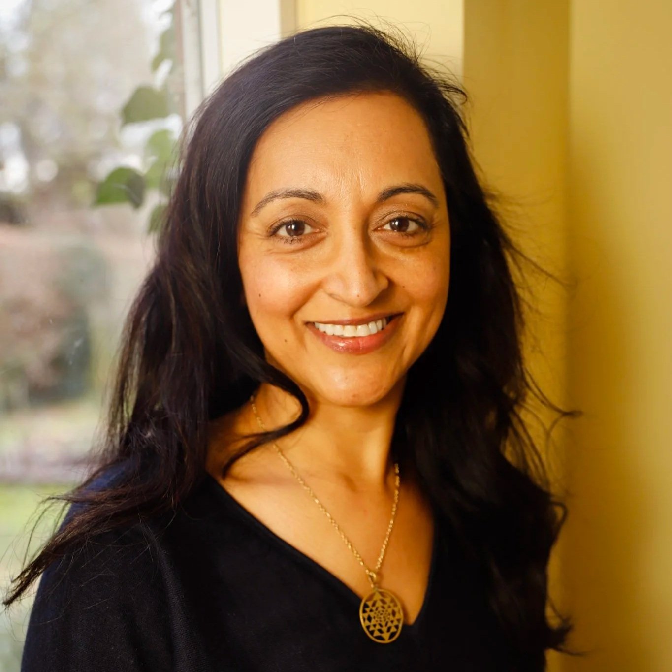 Portrait of Roxana smiling with dark, wavy hair, wearing a black top and a gold necklace, standing near a window with a blurred outdoor background.