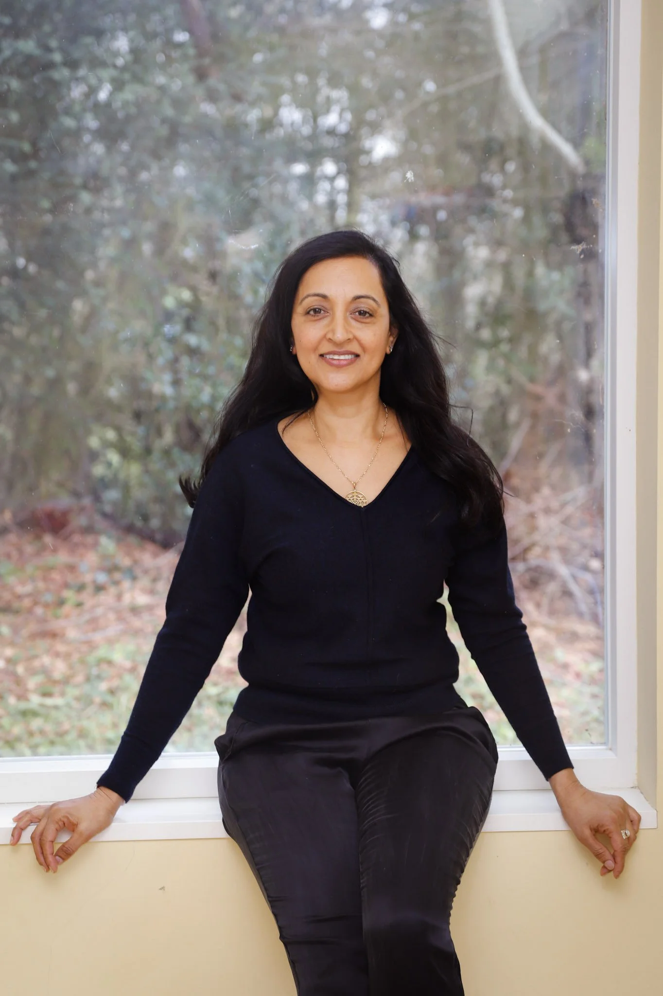 Woman with long dark hair, wearing a black top and pants, sitting on a window sill in front of a large window with a view of trees and leaves outside.