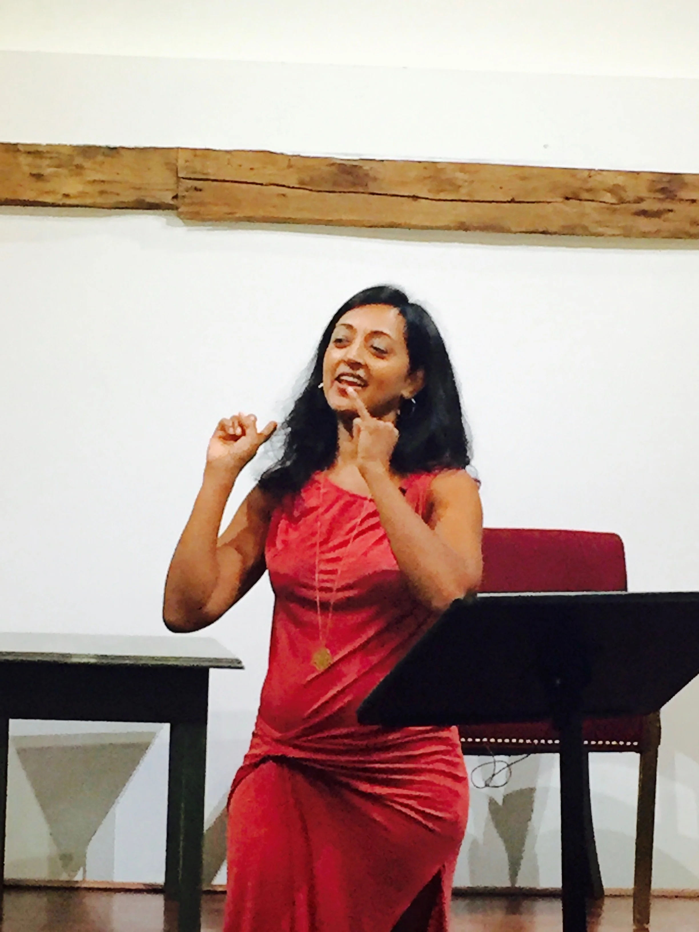 A woman in a red dress standing at a lectern, gesturing with her hands, in a room with white walls and a wooden beam.