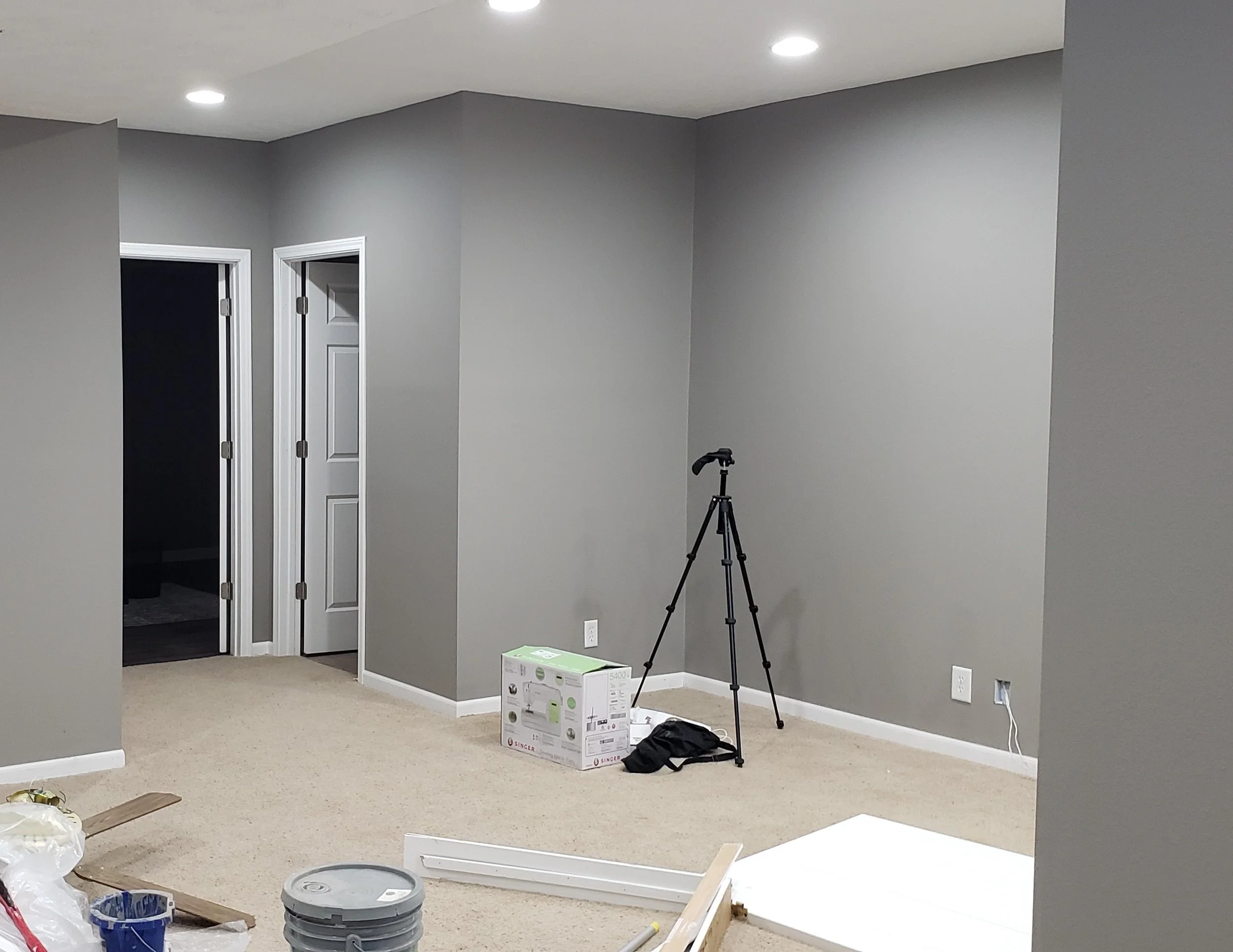 Interior of a room with gray walls and beige carpet, ceiling lights, two open doors leading to dark rooms, a tripod standing near the wall, and construction supplies on the floor.