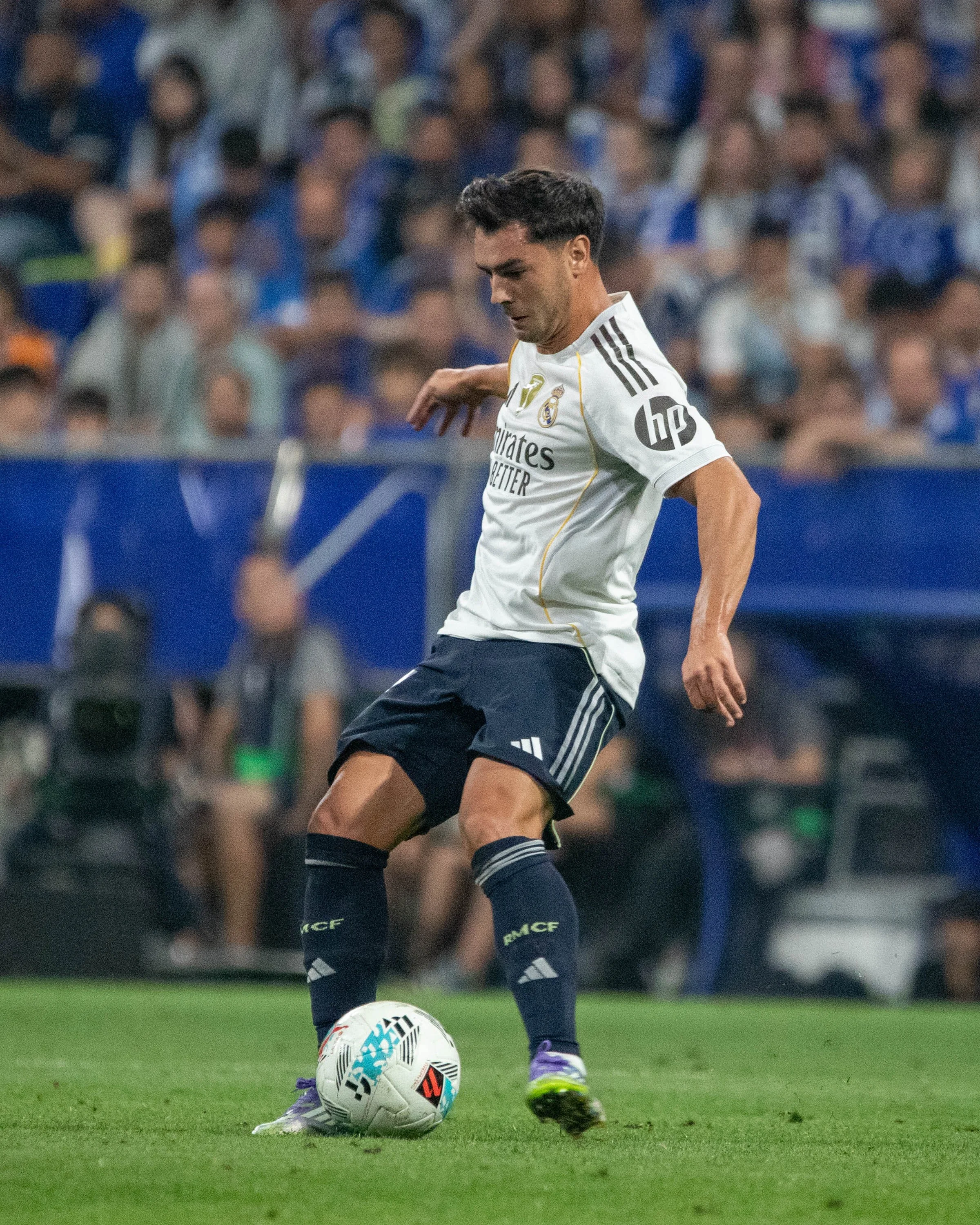 A soccer player in a white and navy uniform with the Real Madrid logo, black shorts, and purple shoes, controlling a soccer ball on the field during a match.
