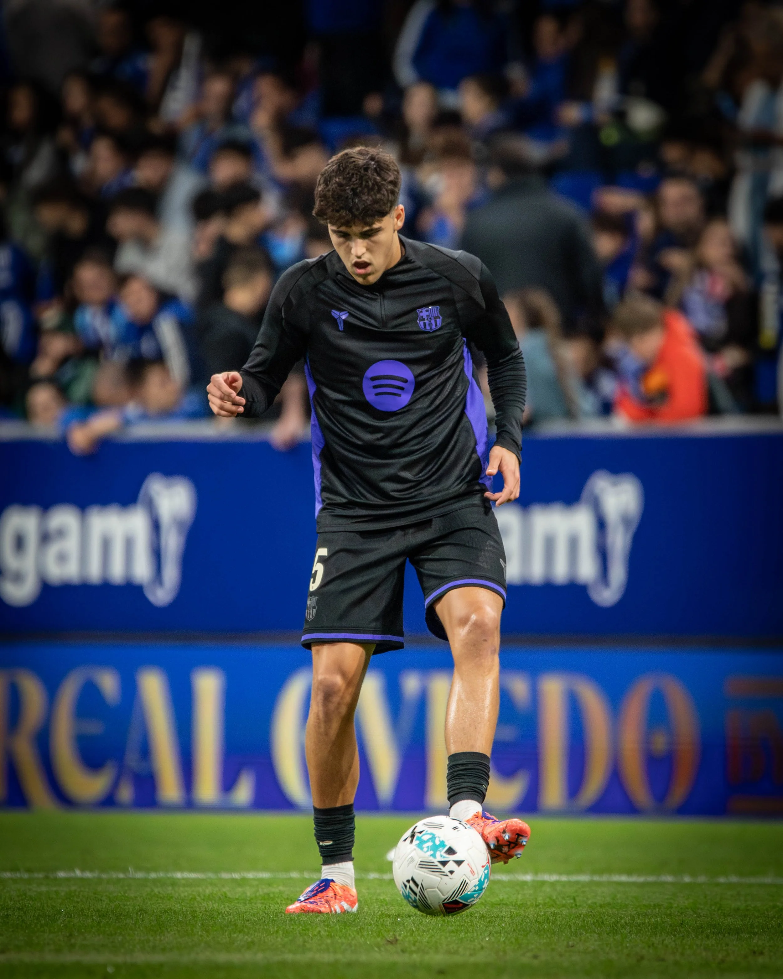 A young male soccer player in black and purple uniform controlling a soccer ball on the field during a match with a crowd in the background.