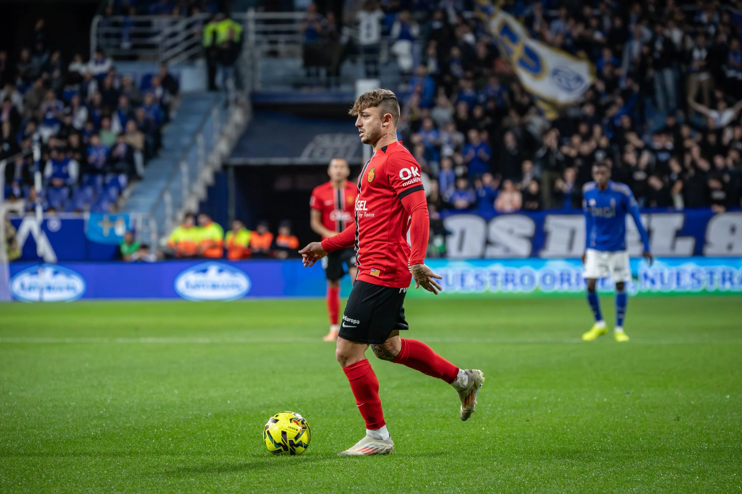 A soccer player in a red jersey and black shorts about to kicks a yellow and black soccer ball during a match, with spectators in the stands in the background.