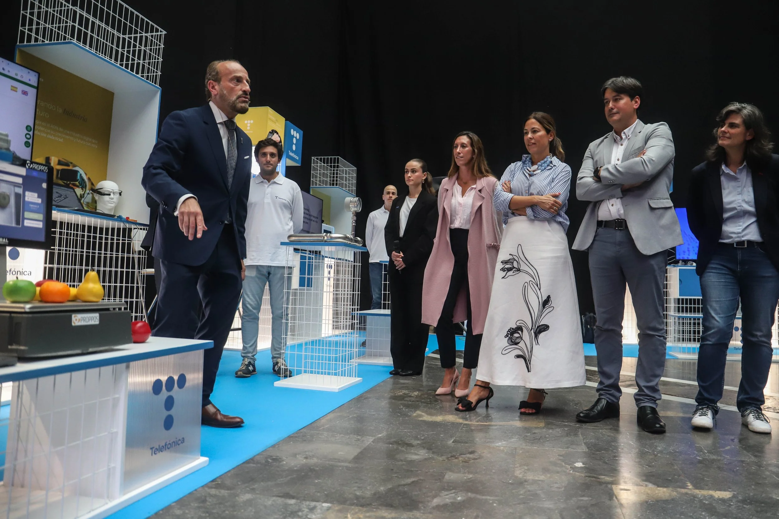 A group of six people, five women and one man, standing in a blue and white exhibit area, listening to a man in a navy suit who appears to be giving a presentation. There are various technological devices and colorful props on display around them.