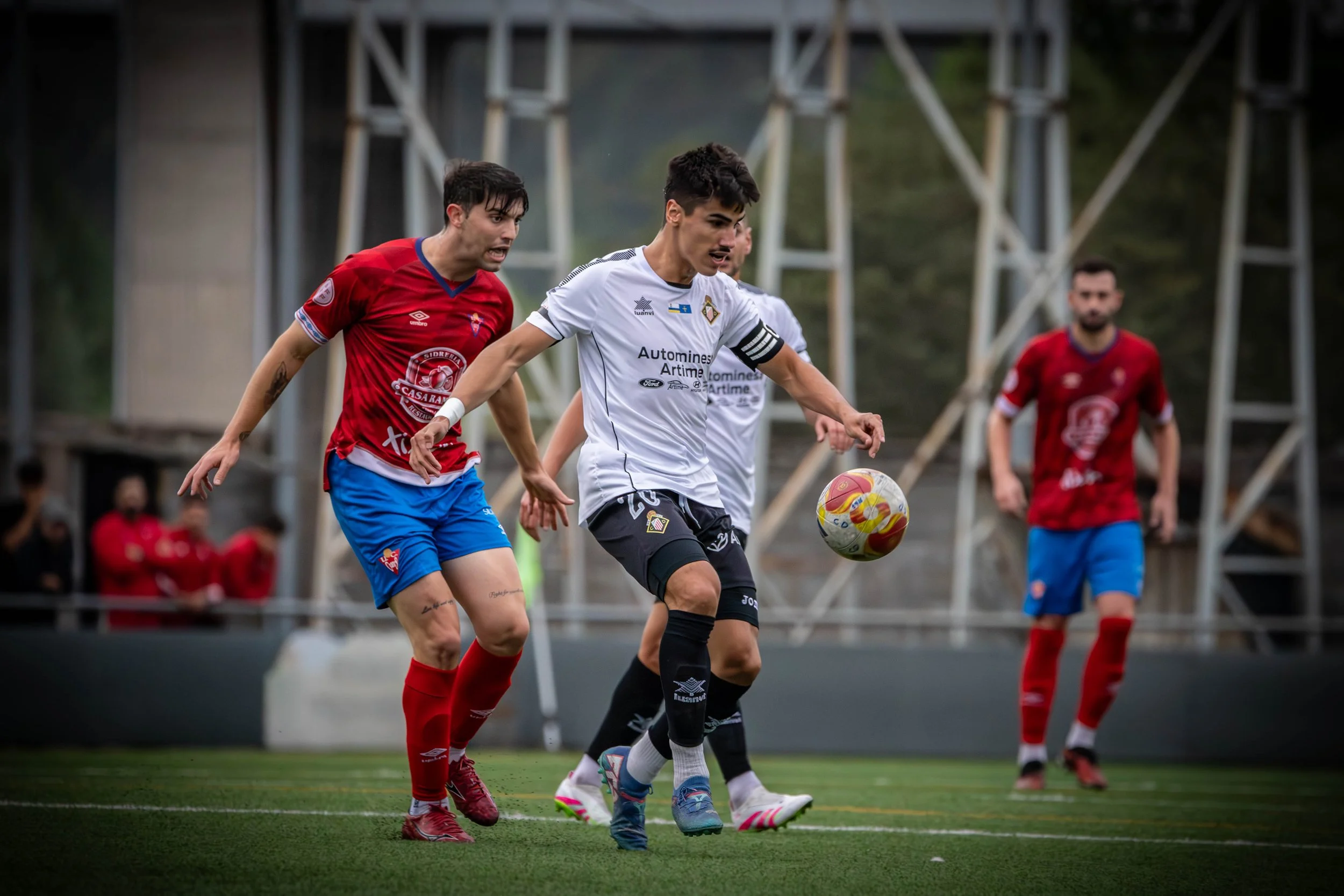 Soccer players competing for the ball on a field, with one player in a white jersey and black shorts controlling the ball, while two players in red jerseys and blue shorts challenge him.