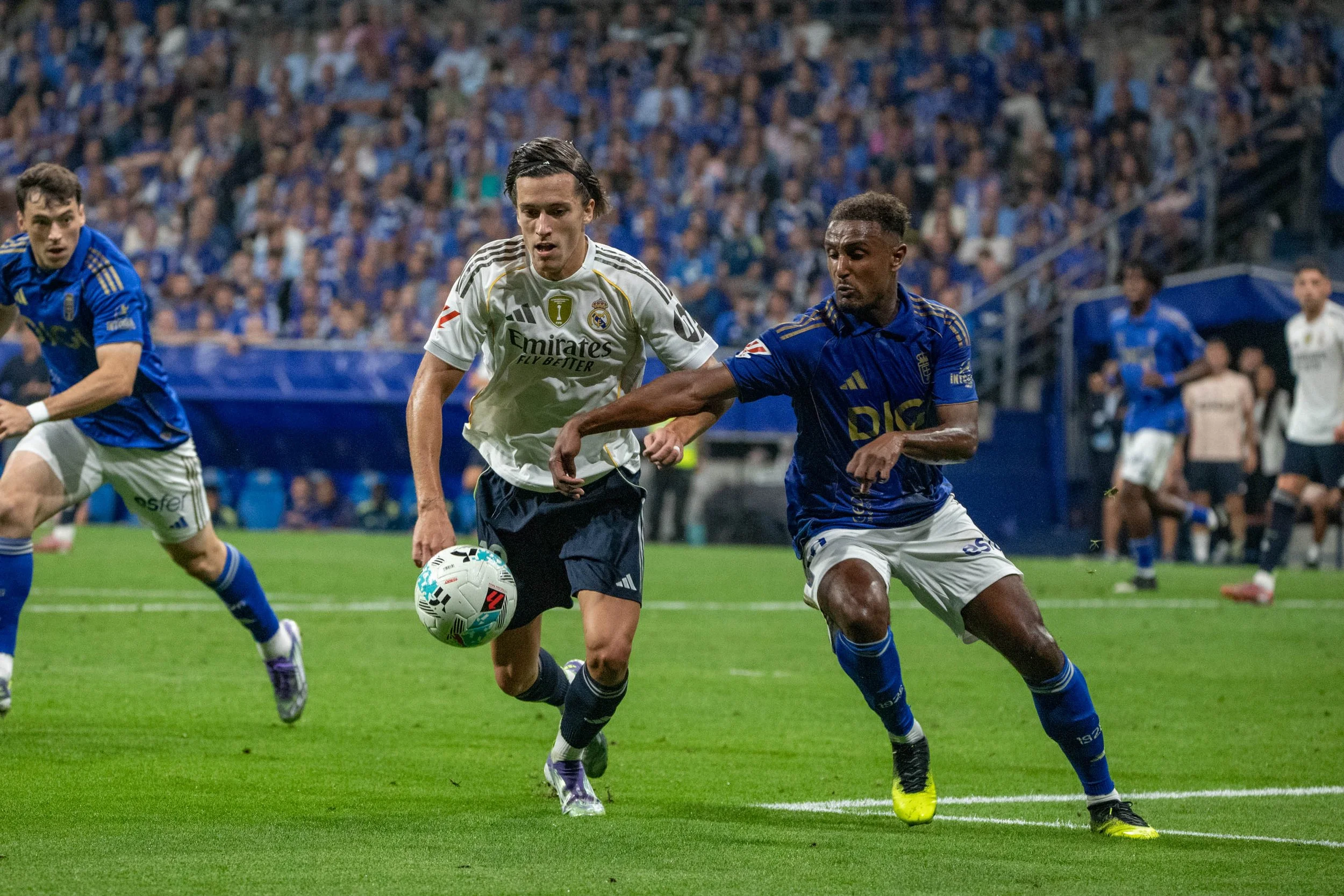 Soccer players in blue and white jerseys chase after the ball on a green field during a game, with a crowd of spectators in the background.