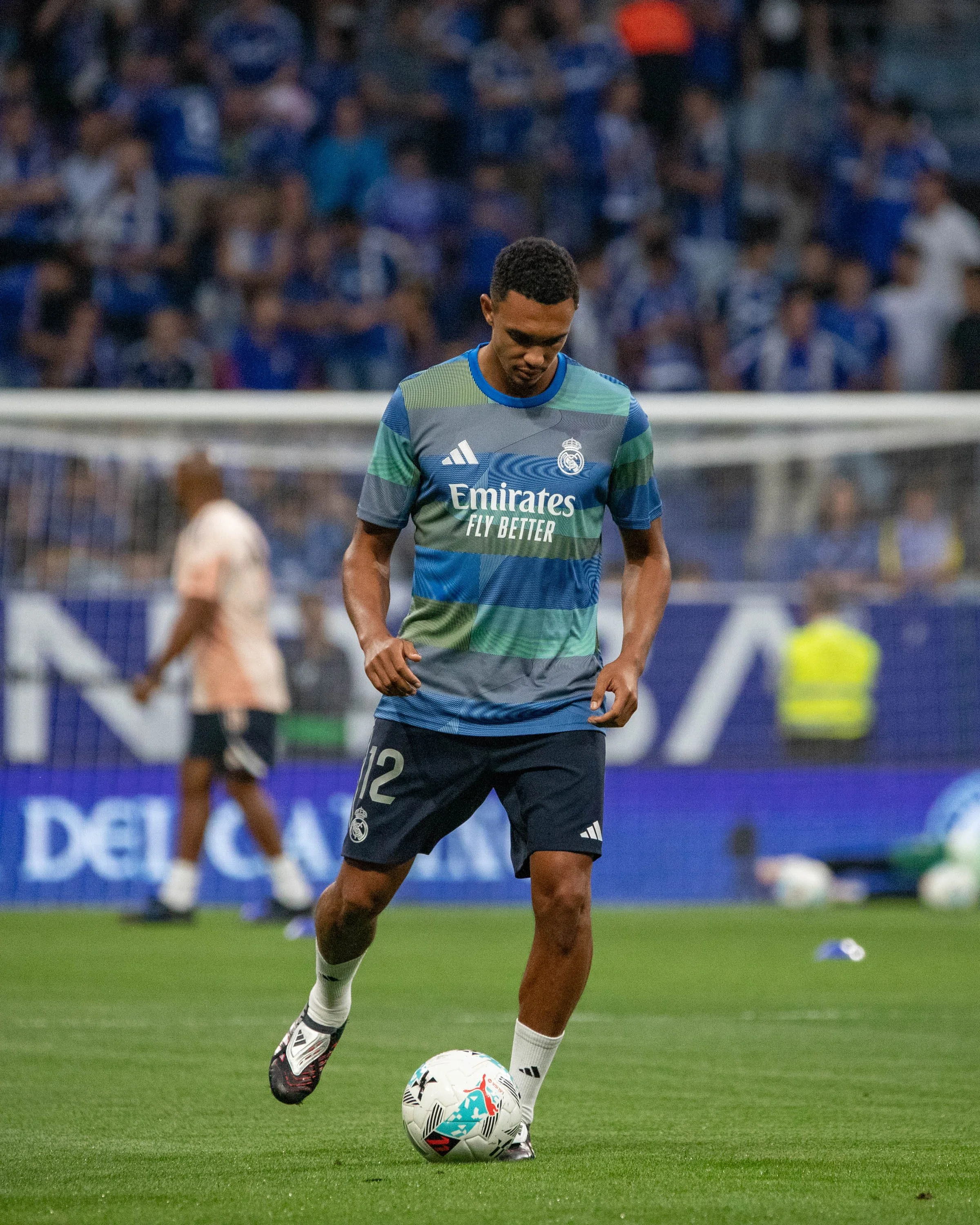 A soccer player in a Real Madrid training jersey kicks a ball on the field during practice or a warm-up session, with a crowd of spectators in the stands behind him.