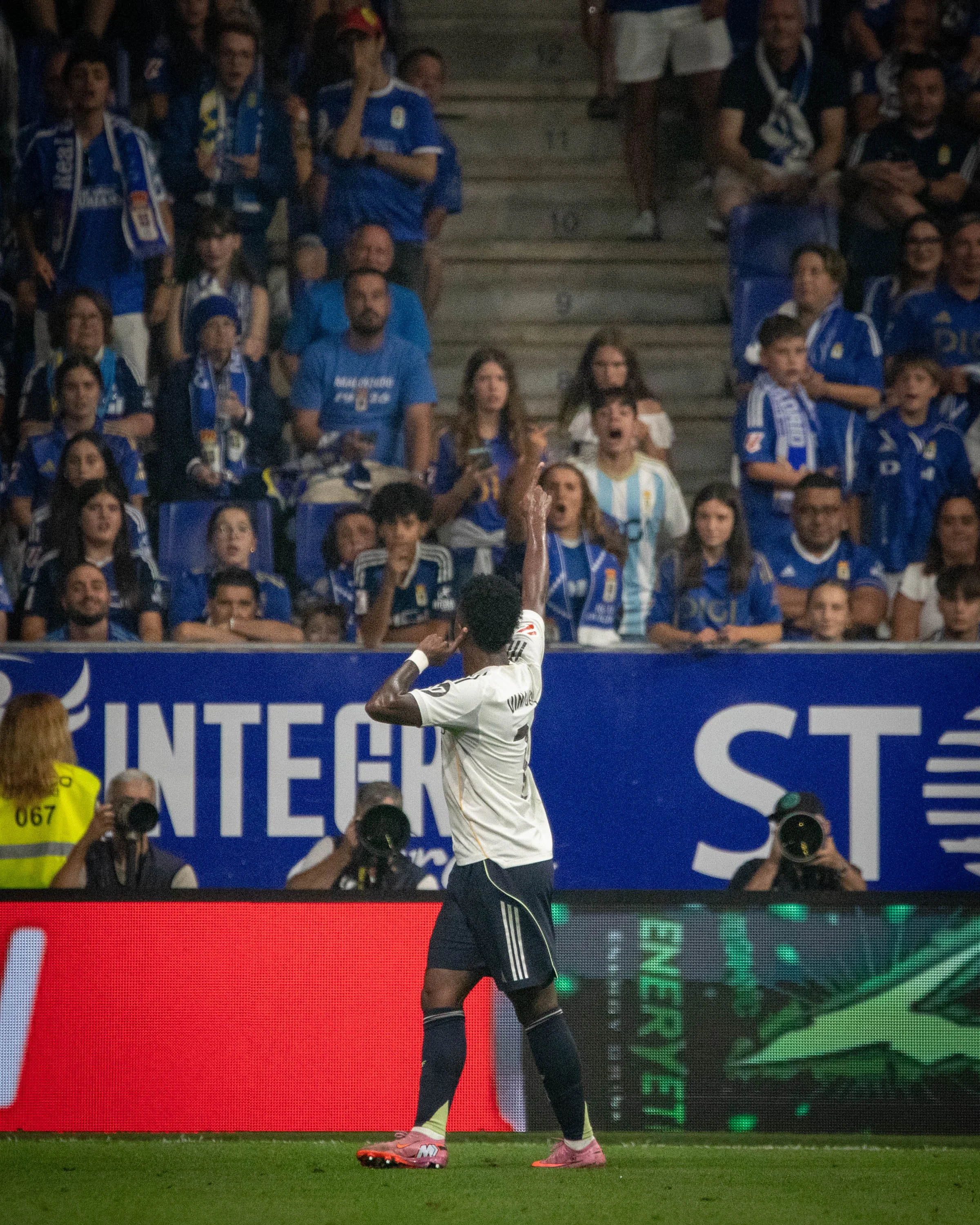 A soccer player in a white jersey and black shorts celebrates after a goal, with fans in blue jerseys and scarves cheering in the stands behind him.