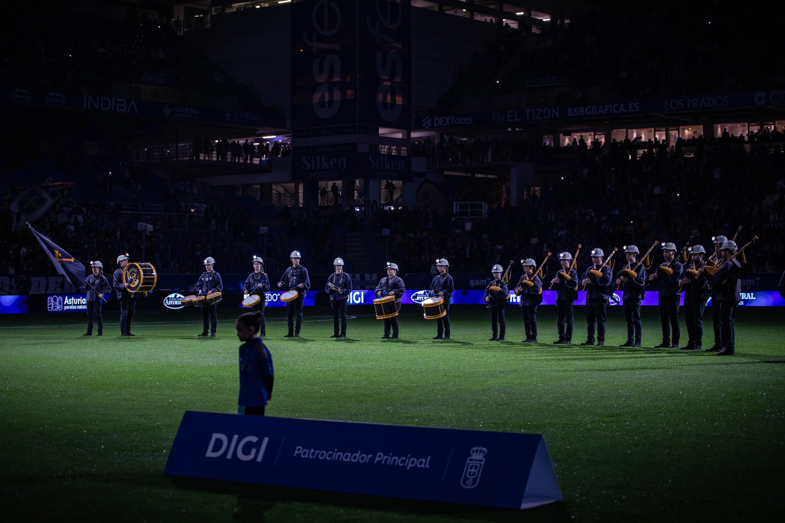 A marching band performing on a football field at night, with members playing drums and wind instruments, and a young child walking in the foreground. The stadium is filled with spectators, and digital advertisements are visible along the sidelines.