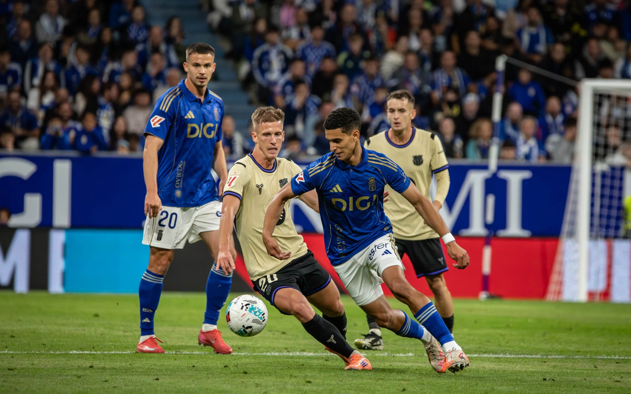 Soccer players competing for ball during match, all in stadium with crowd in background.