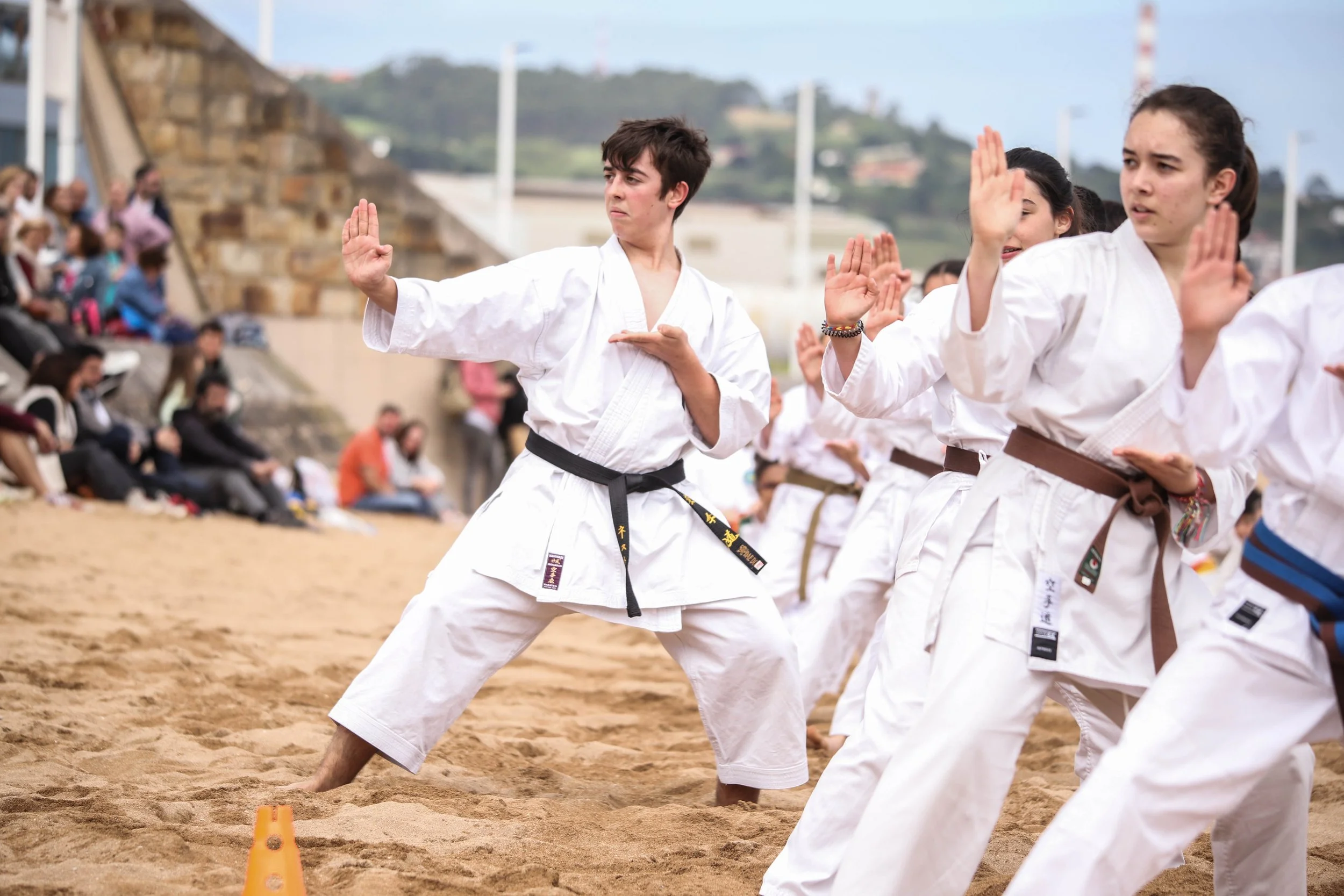 Group of children practicing karate on a sandy outdoor area with spectators in the background.