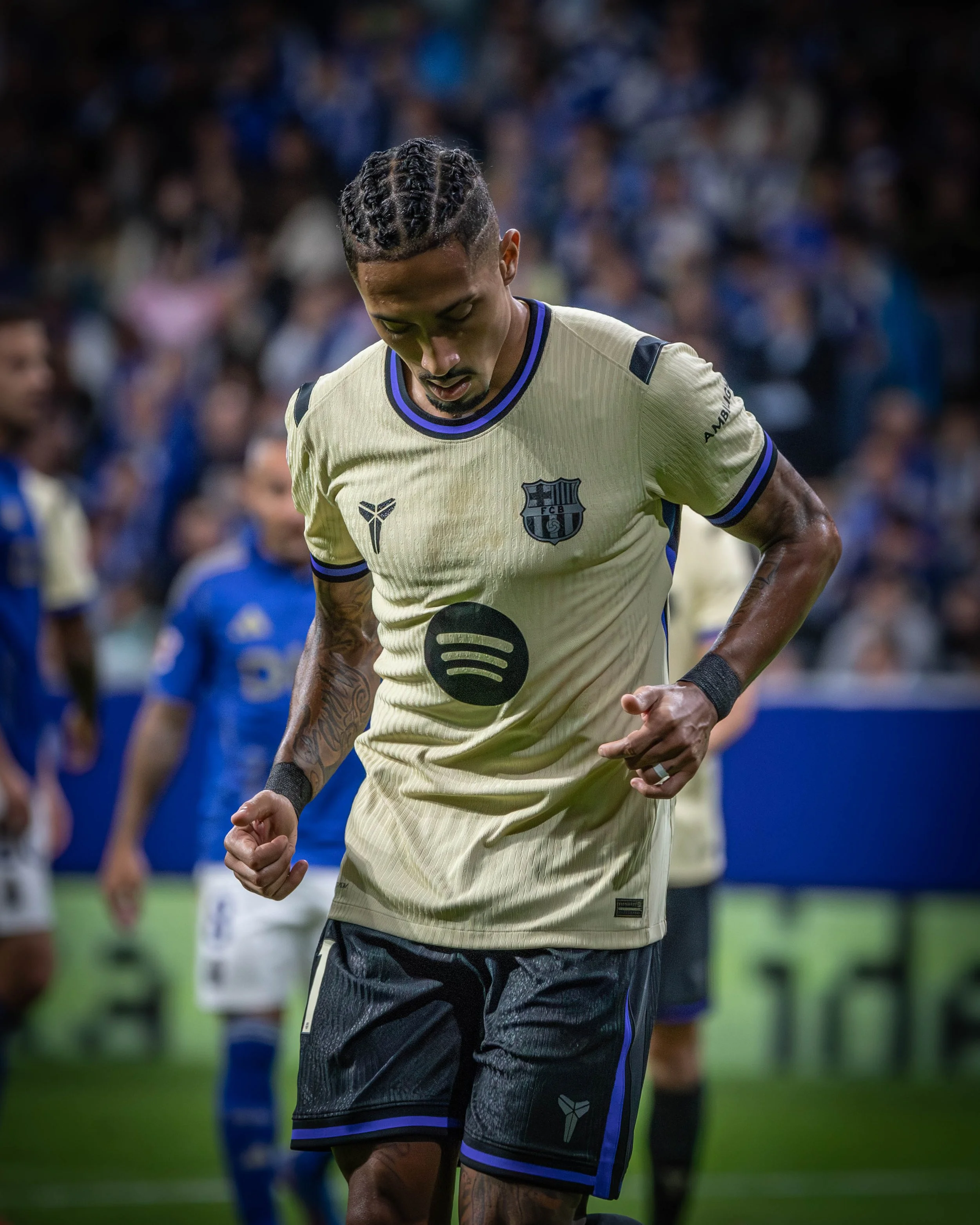 A soccer player wearing a tan and blue jersey with FC Barcelona's crest and the Spotify logo, walking on the field during a match.