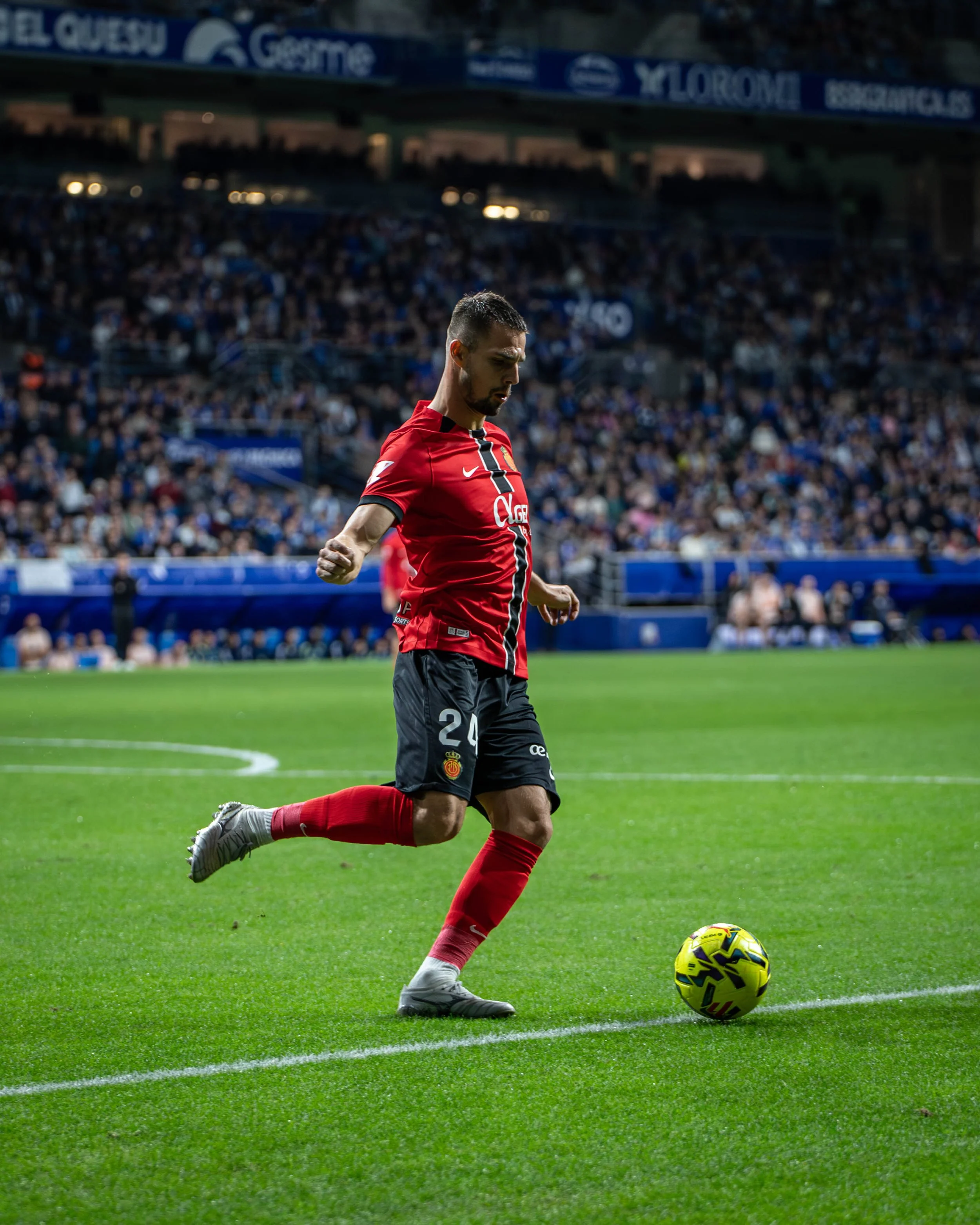 A soccer player in a red and black uniform is about to kick a yellow soccer ball on the field with a crowd in the stands behind him.
