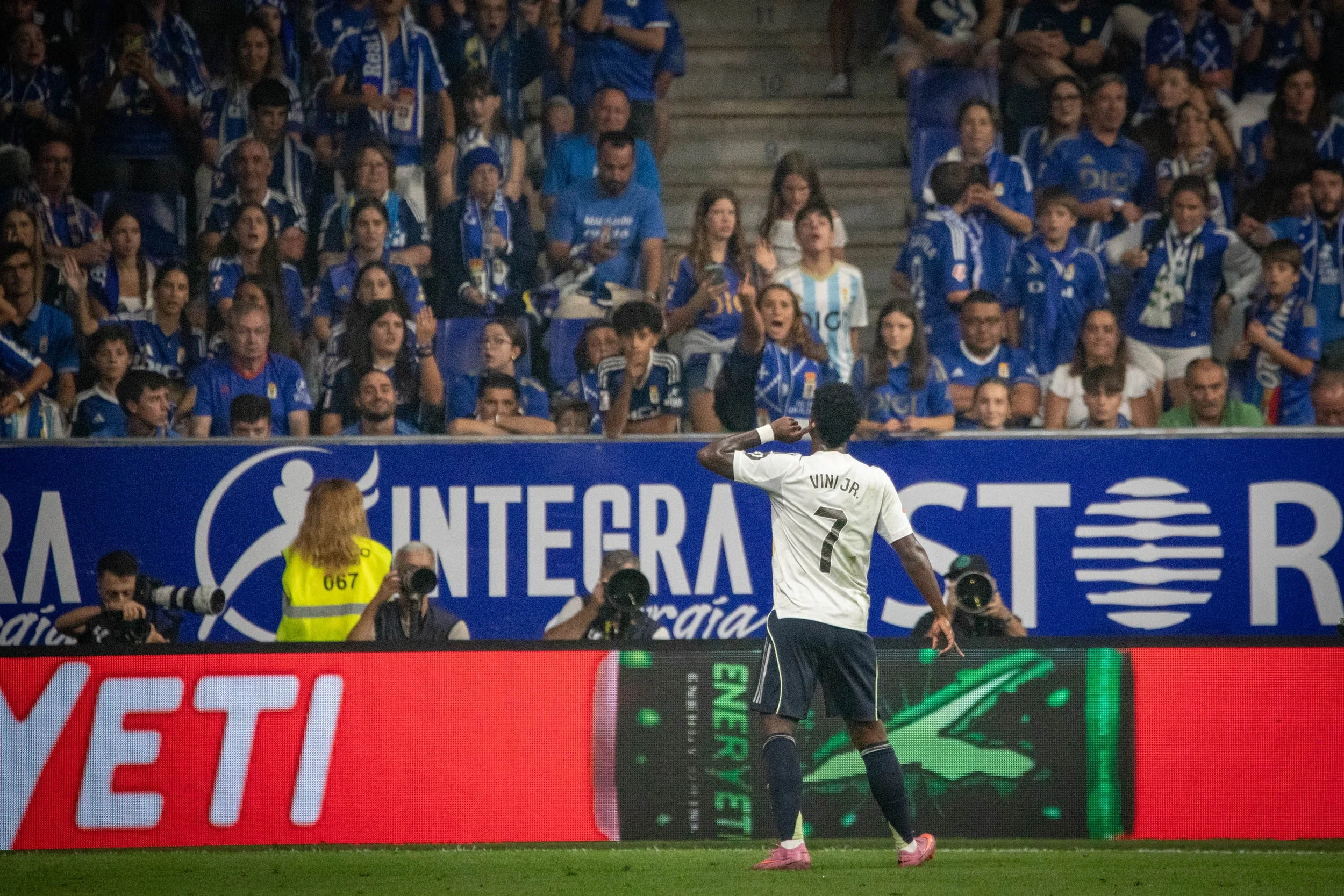 A soccer player wearing a white jersey with the name 'Vini Jr.' and the number 7 is celebrating on the field, with a large crowd of fans in blue jerseys cheering behind a barrier adorned with advertising.