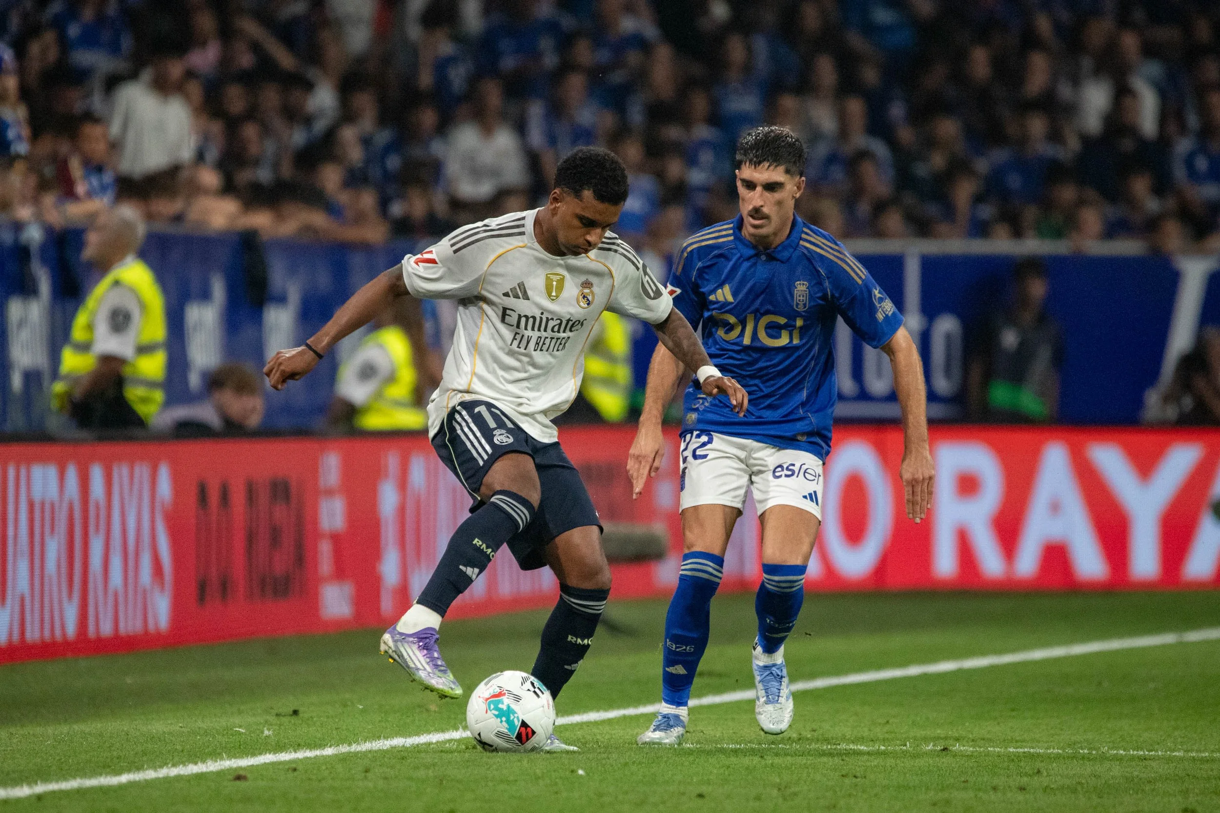 Two soccer players, one in a white and navy uniform and the other in a blue uniform, compete for the ball on the field during a match with spectators in the background.