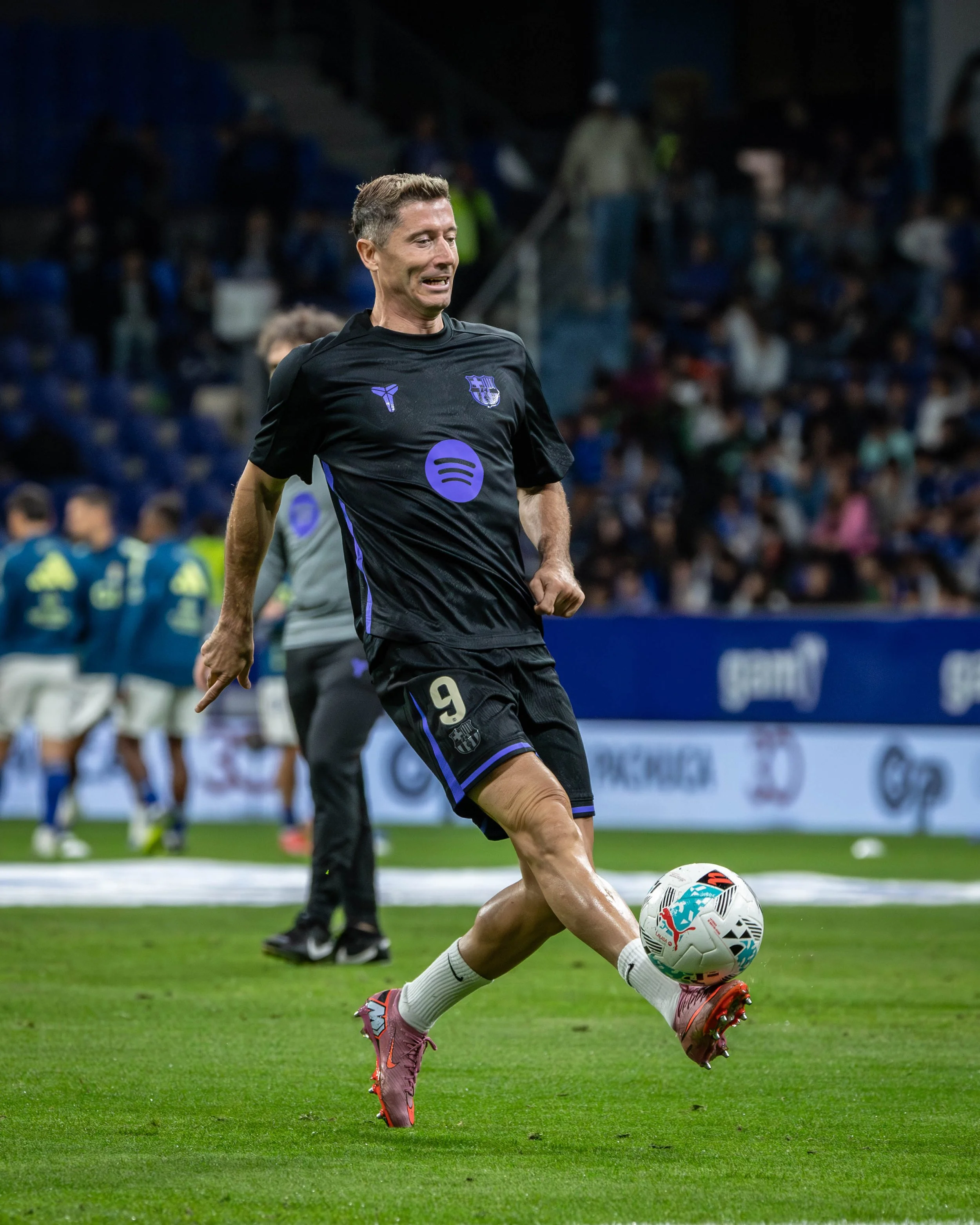 A soccer player wearing a black jersey with the number 9, kicking a soccer ball on the field during a game at night.
