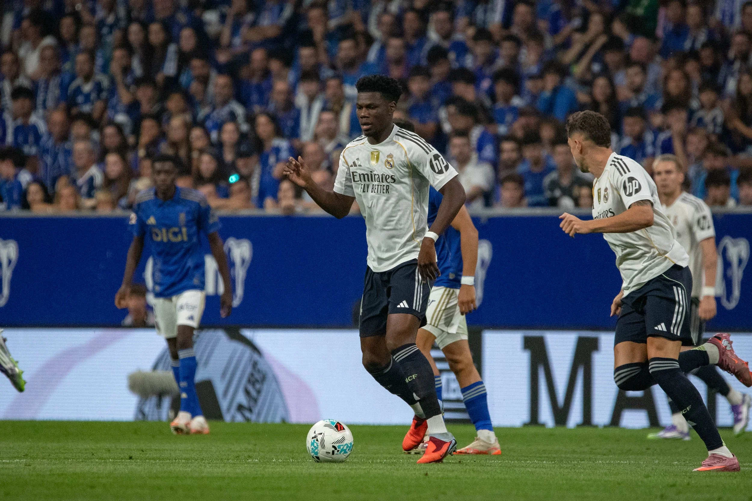 Soccer players in white and blue jerseys during a match, with a player in a white jersey at the center dribbling the ball on the field with an audience watching from the stands.