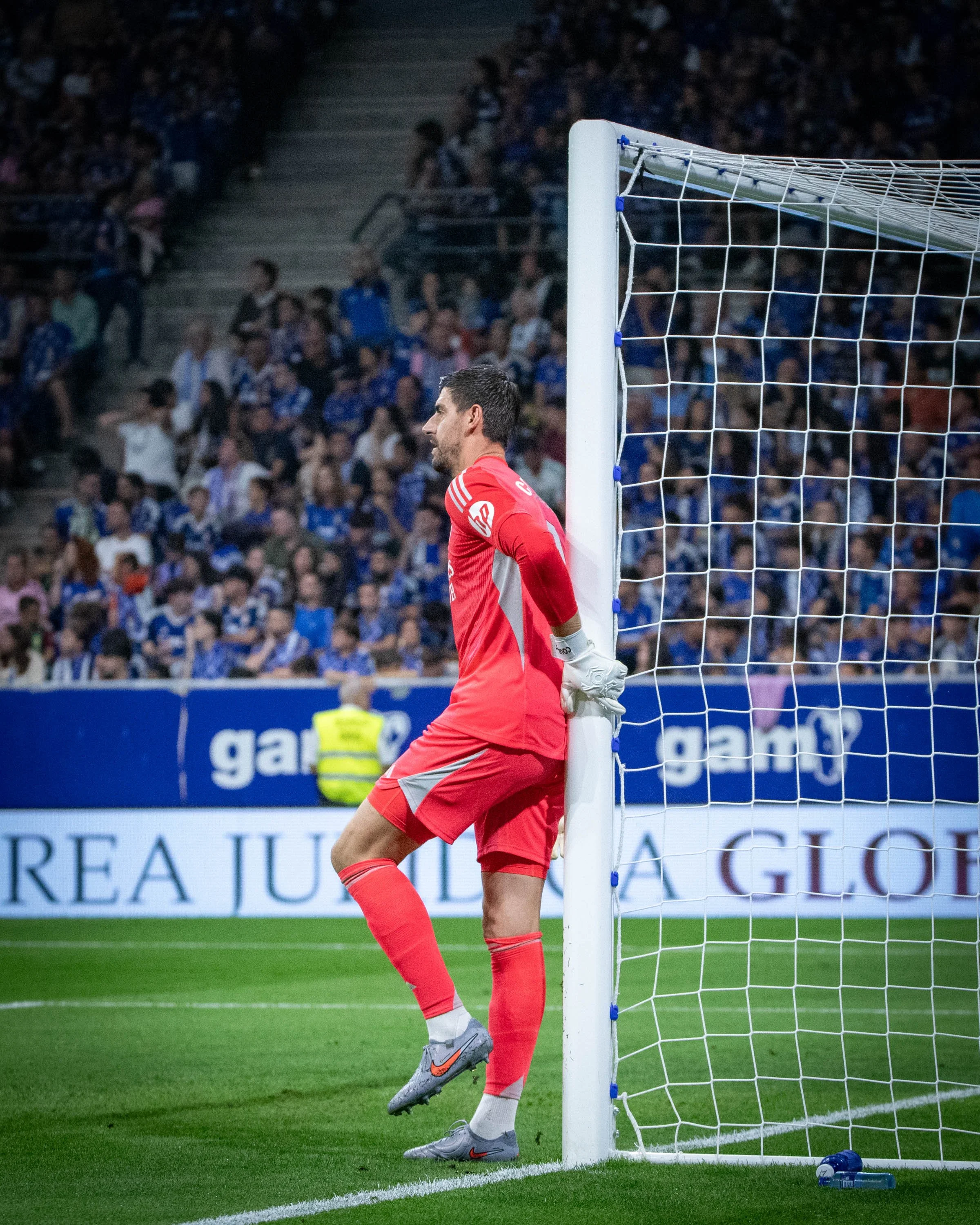 A soccer goalkeeper in red uniform leaning against the goalpost on a soccer field during a match, with a large crowd in the background.