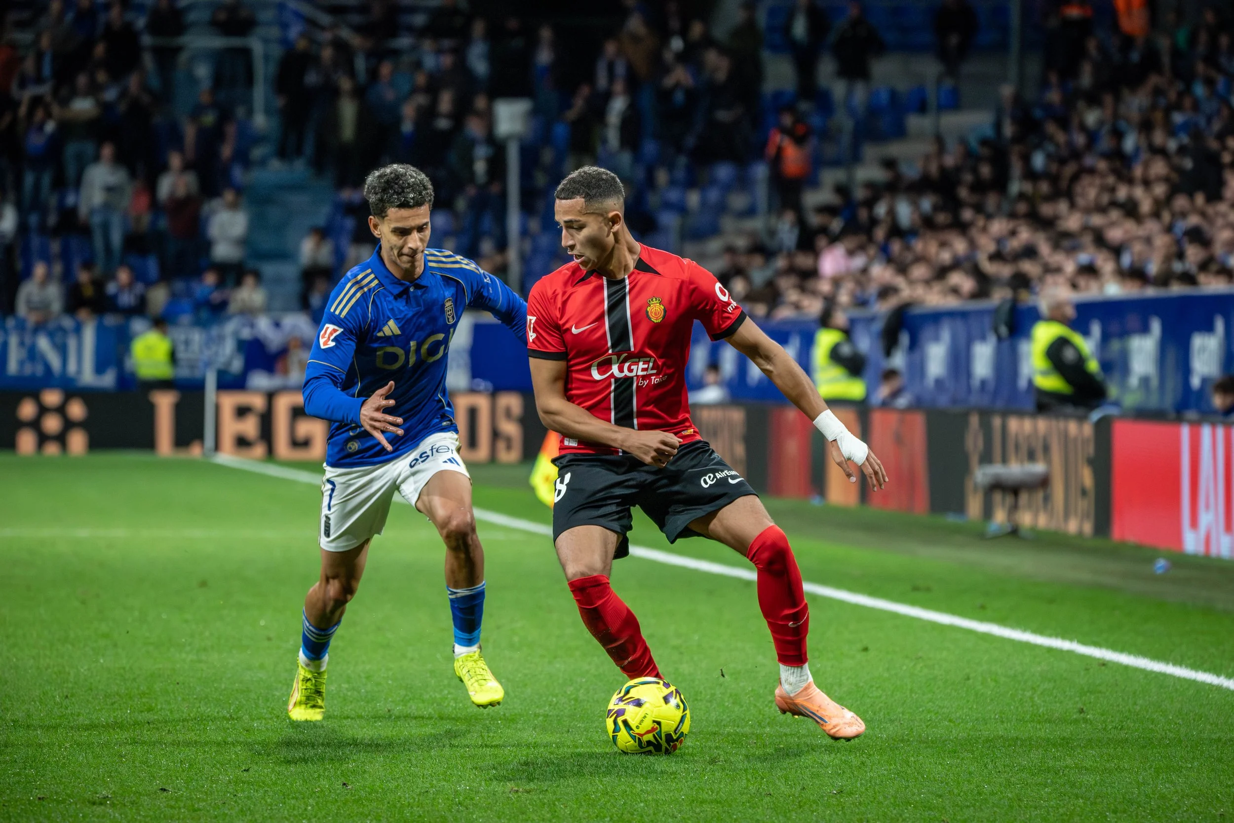 Two soccer players chase the ball on the field during a match, with a crowd in the stands watching.