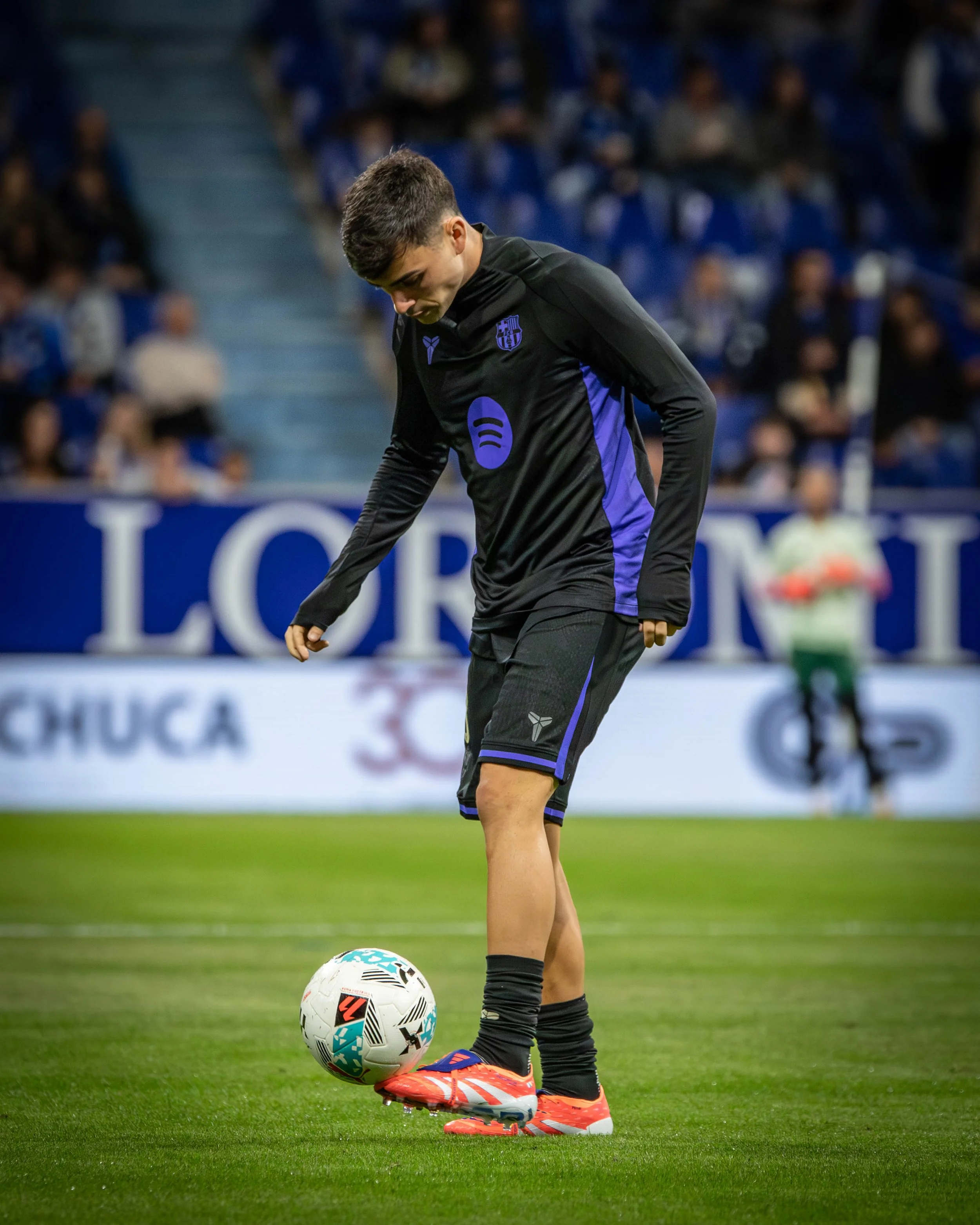 A male soccer player in black and blue uniform preparing to kick a soccer ball on the field with spectators watching in the background.