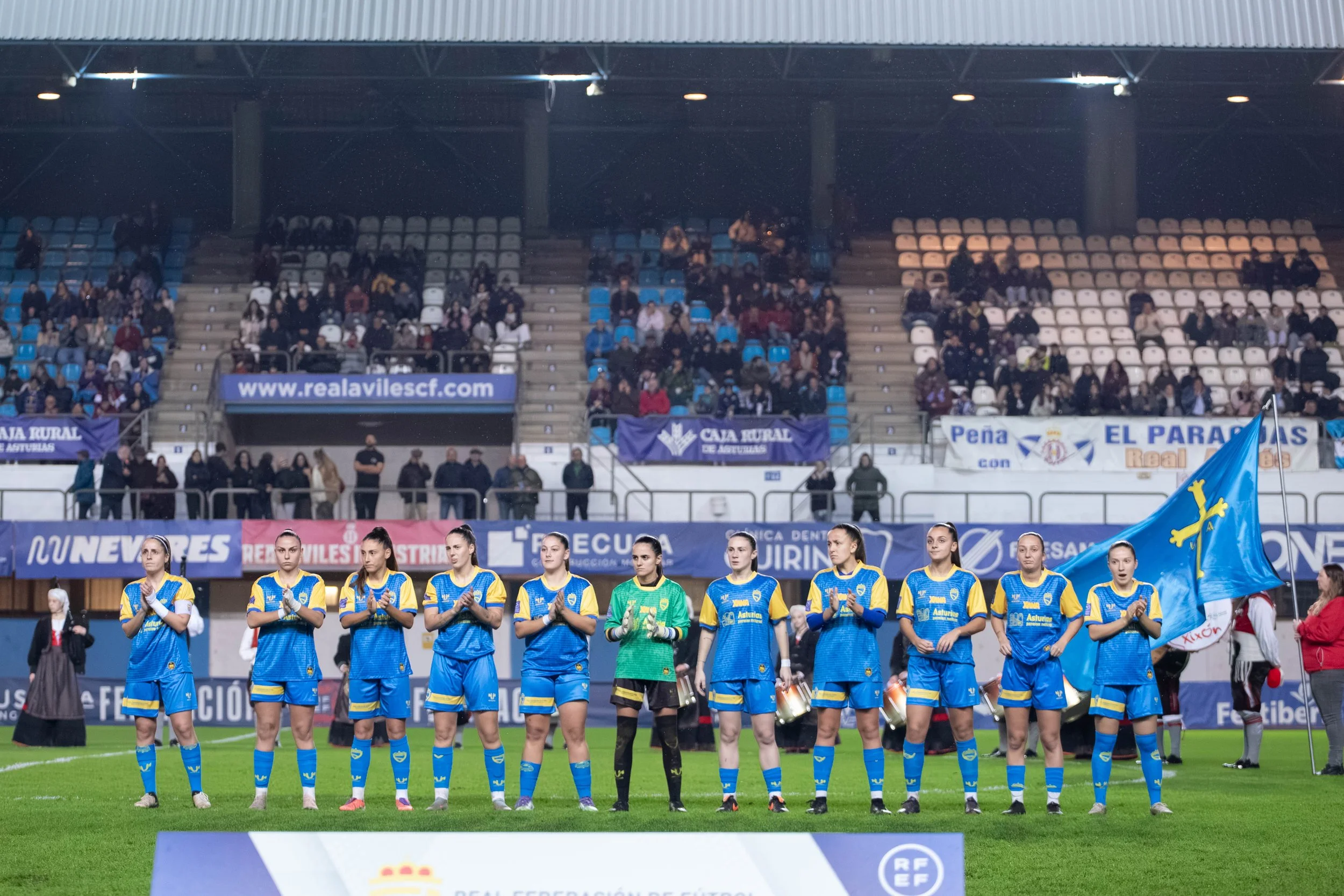 A women's soccer team standing on a field in blue and yellow uniforms, hearing before a game or ceremony at a stadium with spectators in the stands.