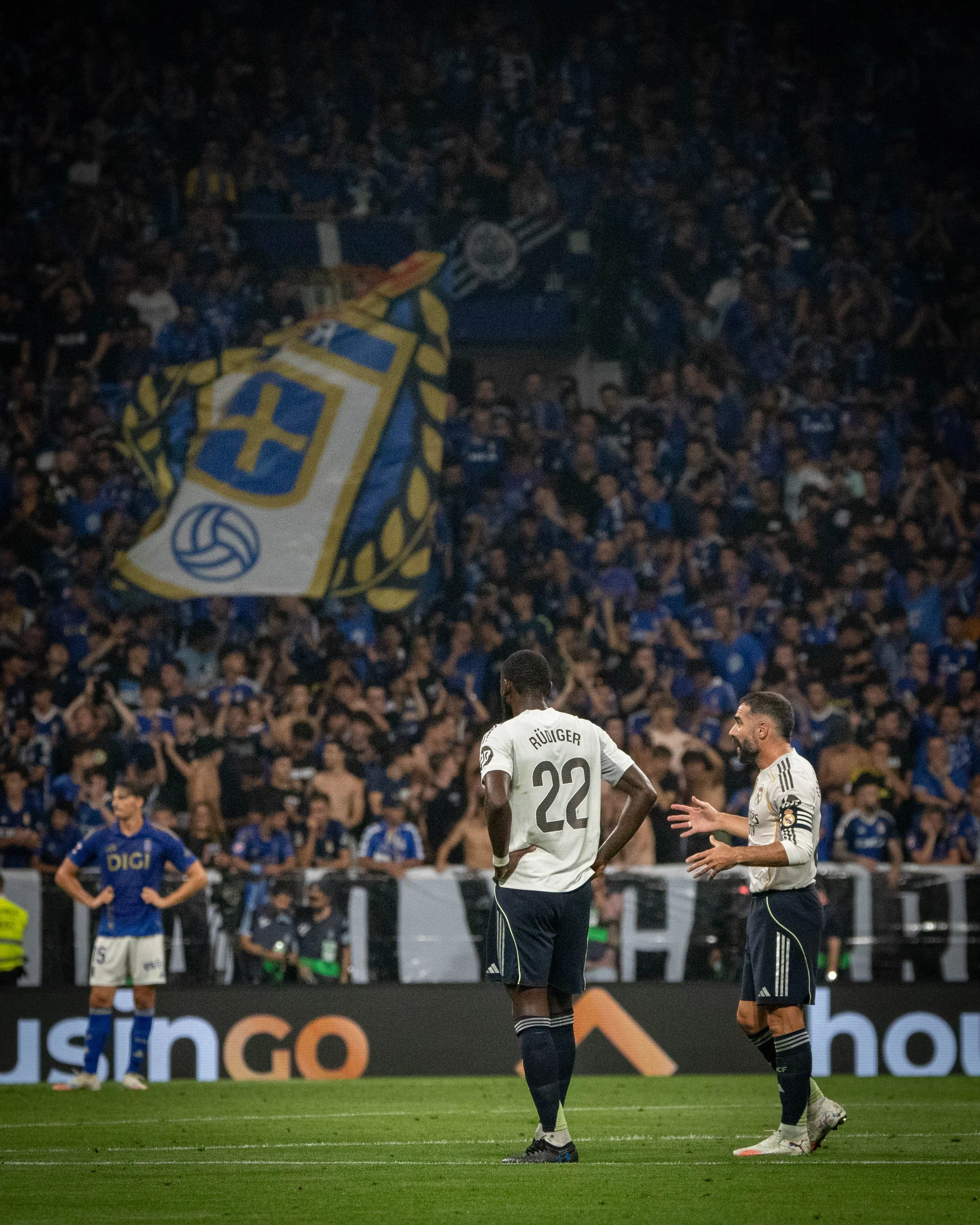 Two soccer players on the field, one wearing a white jersey with number 22 and the other wearing a white jersey with captain's armband, engaging in a conversation during a match. In the background, there is a crowd of fans holding a large flag with a
