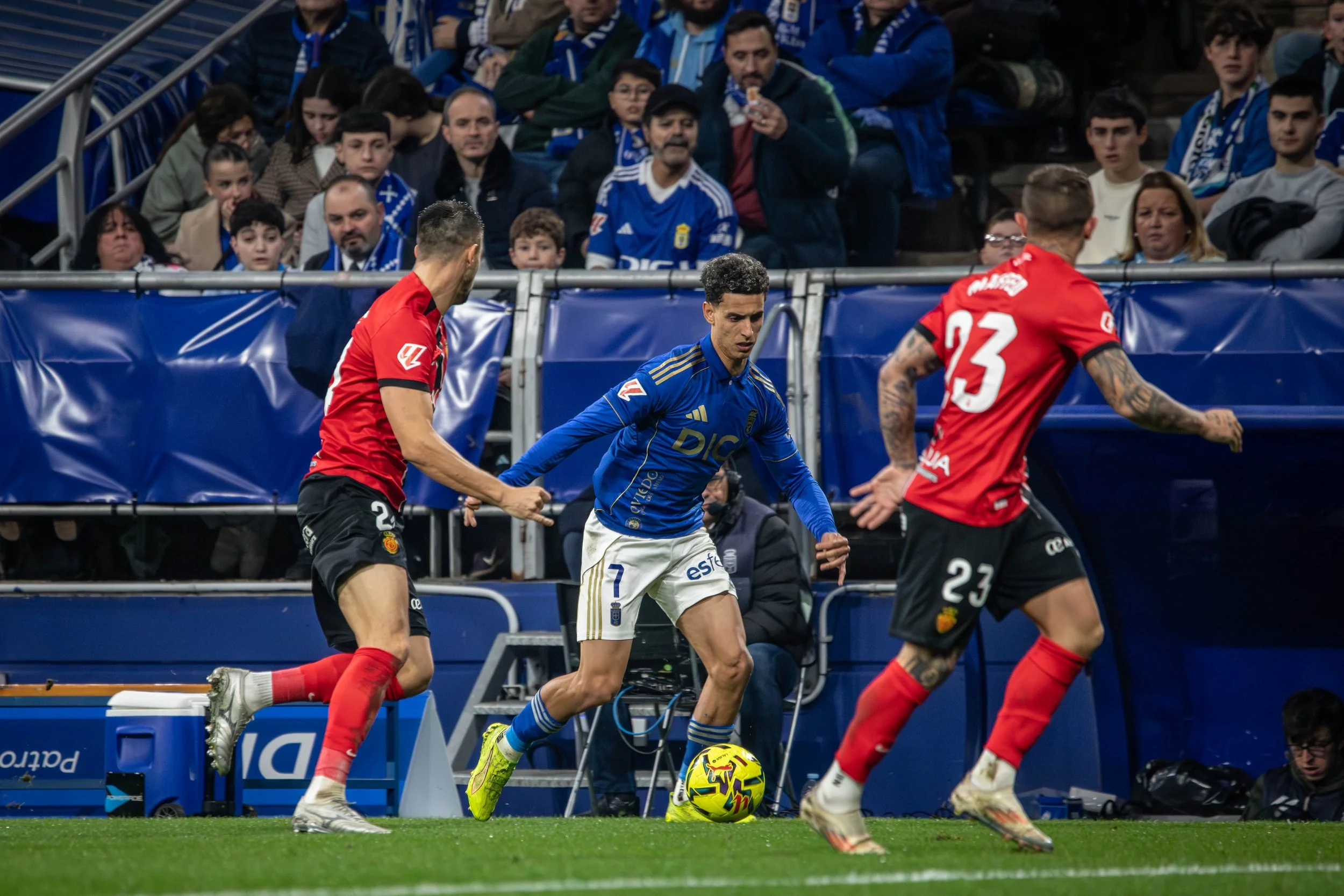 A soccer player in a blue jersey with the number 7 is controlling the ball near the sideline while two players in red jerseys with numbers 2 and 23 are approaching from behind during a match in a stadium with spectators watching.