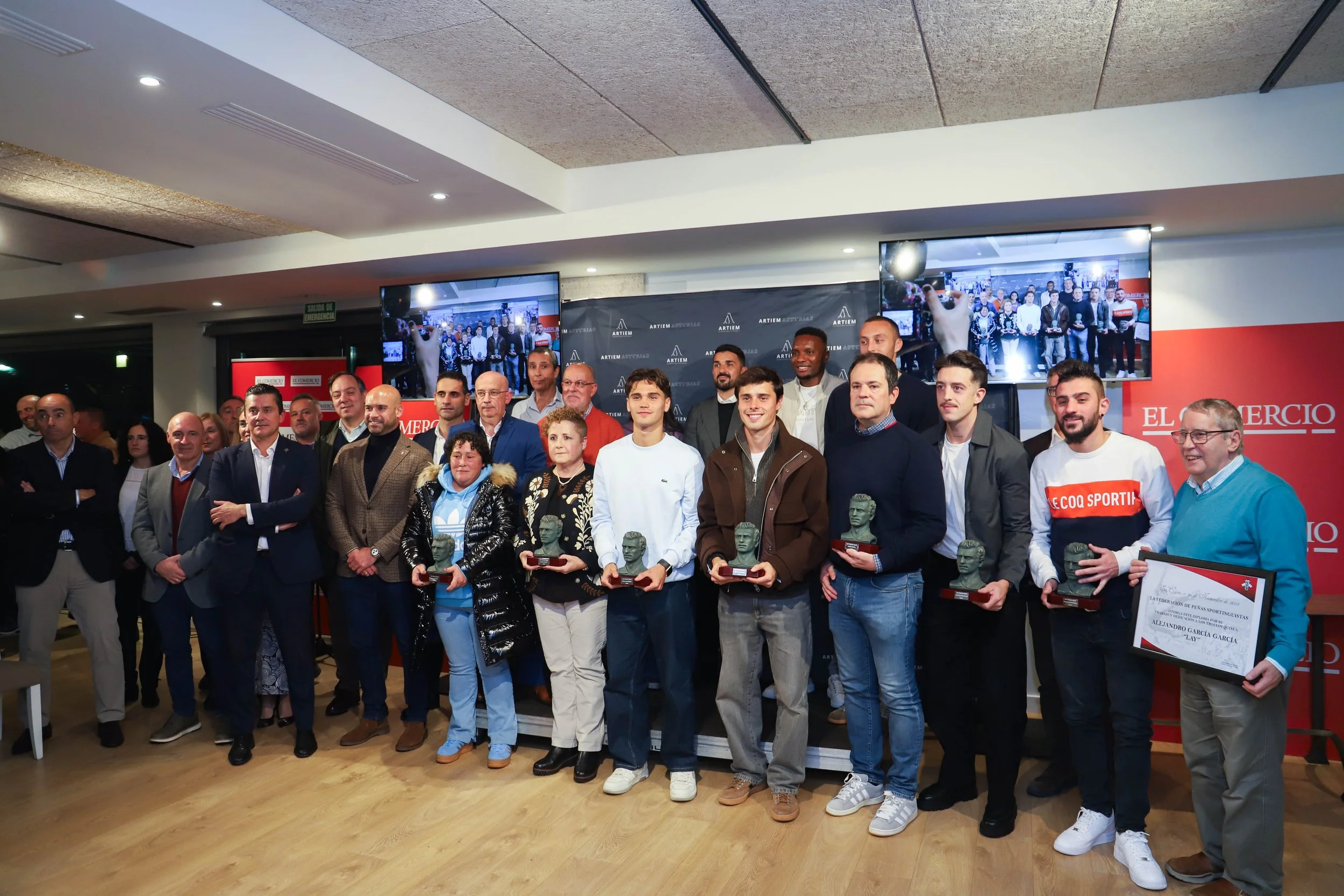 Group of people at an award ceremony, holding trophies and framed certificates, standing on a stage with a backdrop and television screens displaying a group photo.