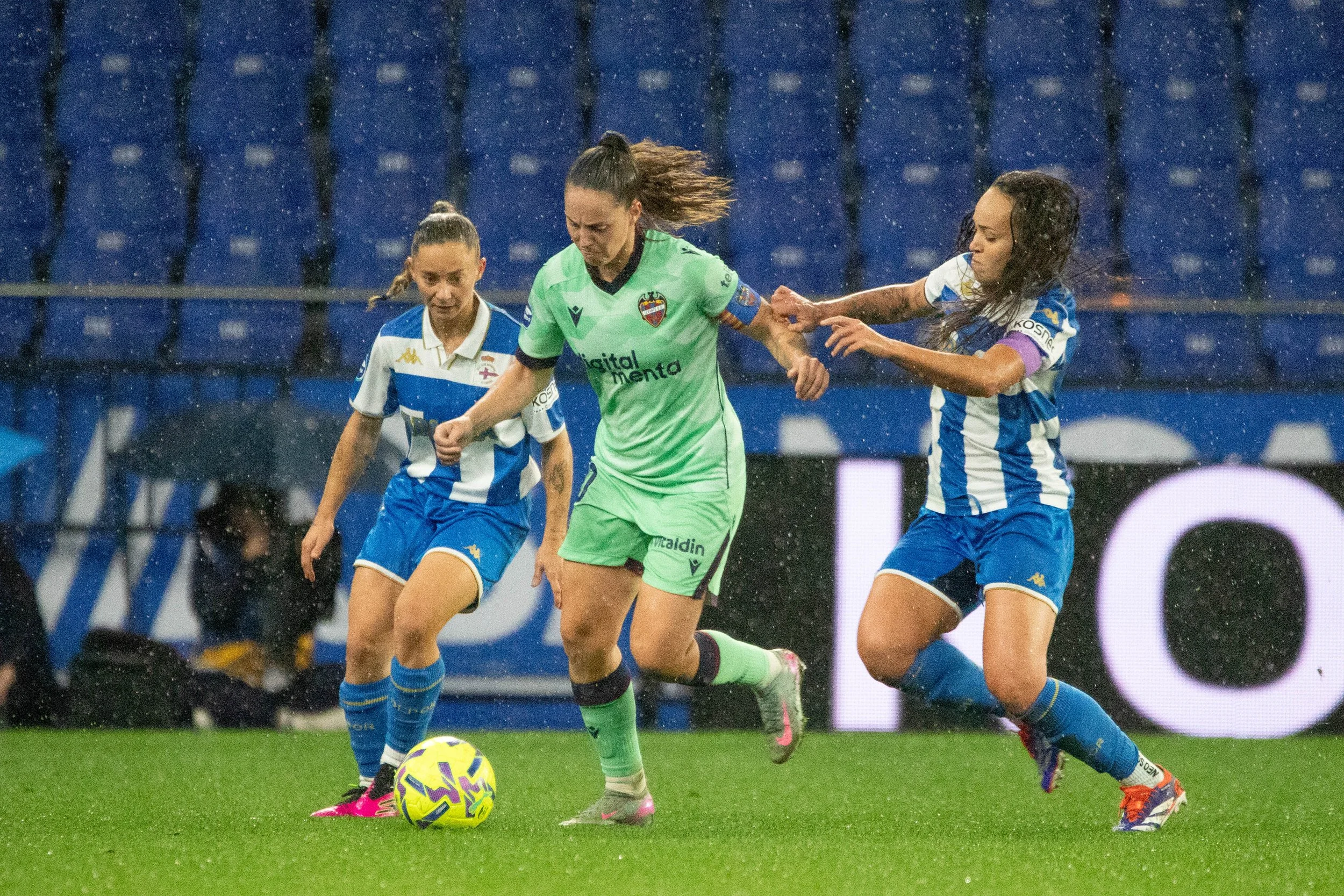 Three female soccer players competing for the ball during a match in rainy weather, with blue stadium seats in the background.