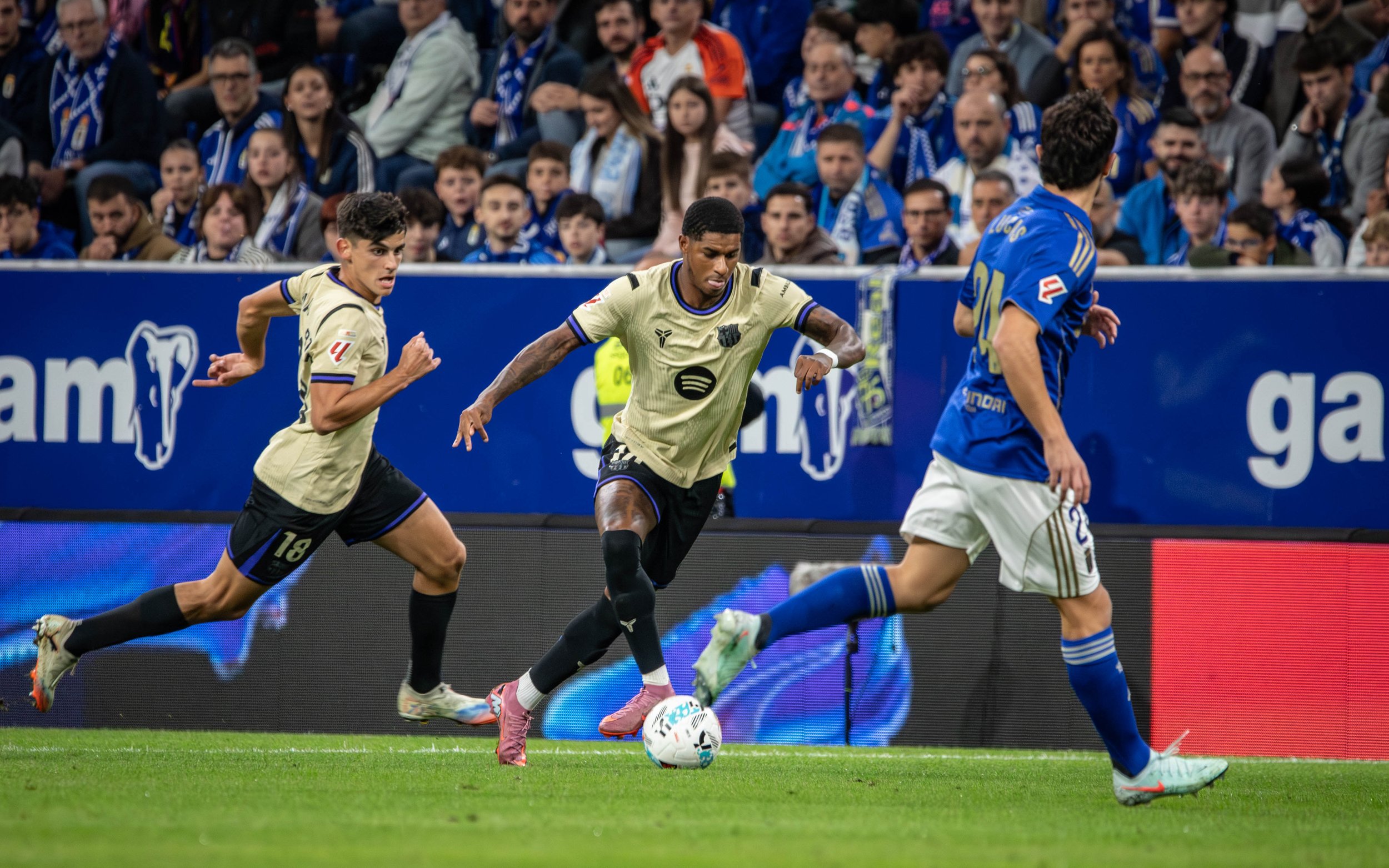 Three soccer players competing for the ball during a match, with a crowd of spectators in the background.