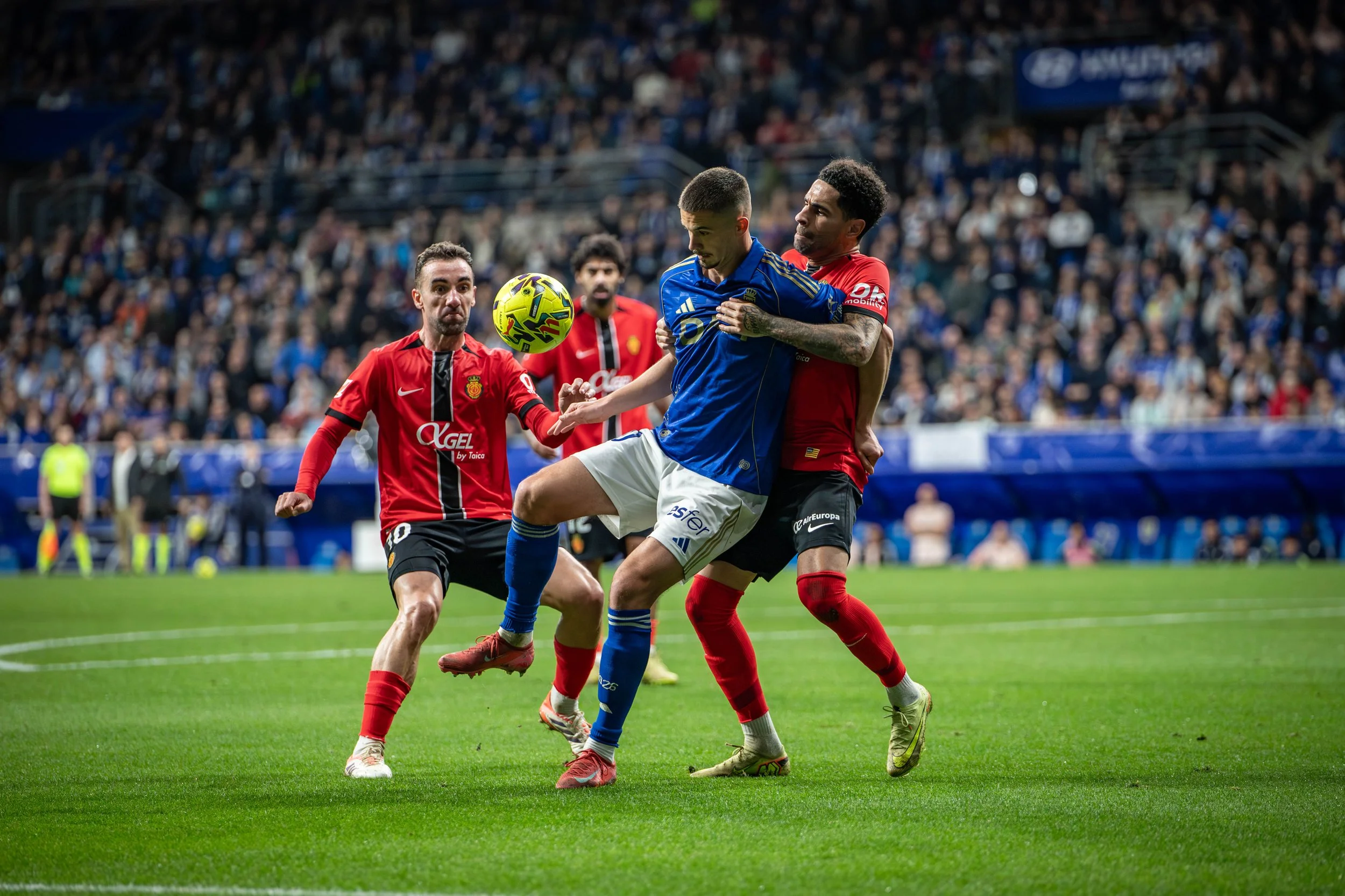 Soccer players in red and blue jerseys competing for ball on field during match with crowd spectators in background.