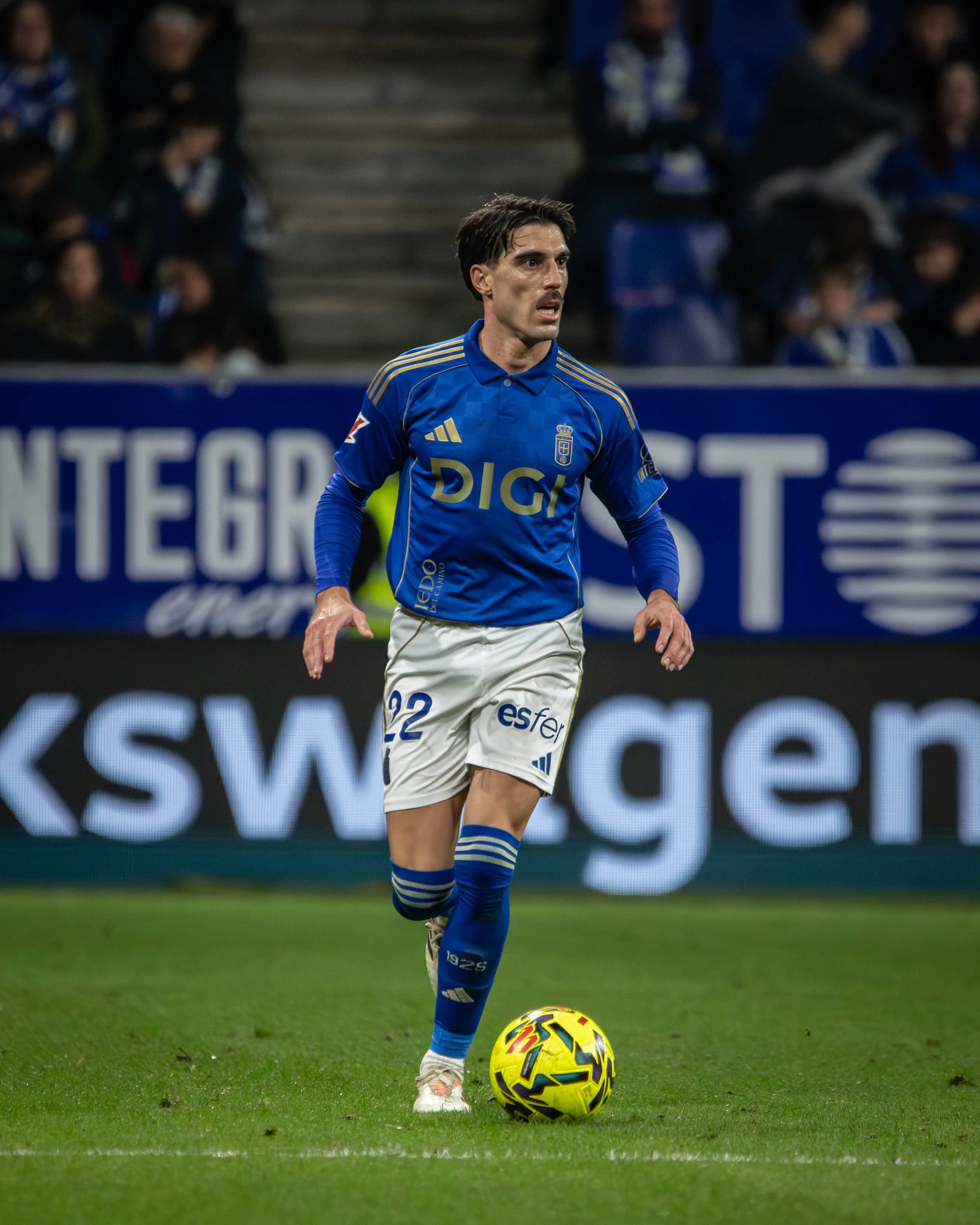 A soccer player in a blue jersey and white shorts dribbling a yellow and purple soccer ball on the field during a match.