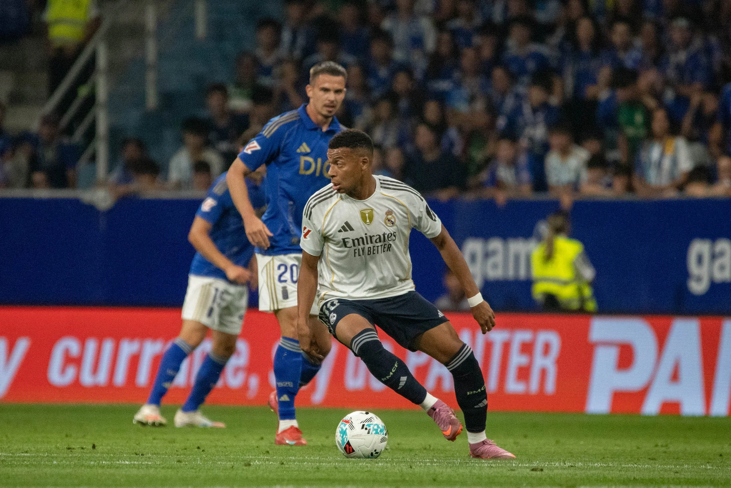 Soccer players from Real Madrid and another team competing for ball on field with crowd in stands.