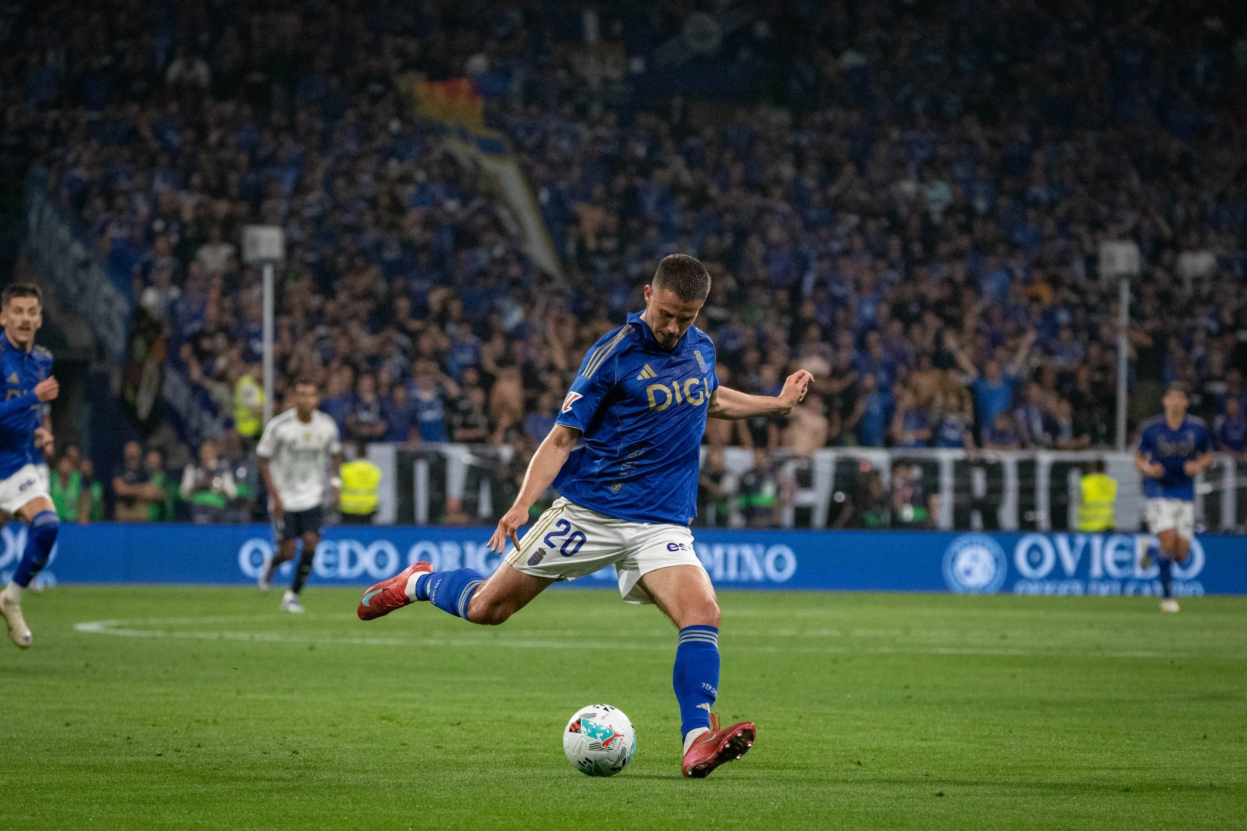 A soccer player in a blue uniform with the number 20 kicks a white soccer ball on a green field during a match, with a crowd in the background.