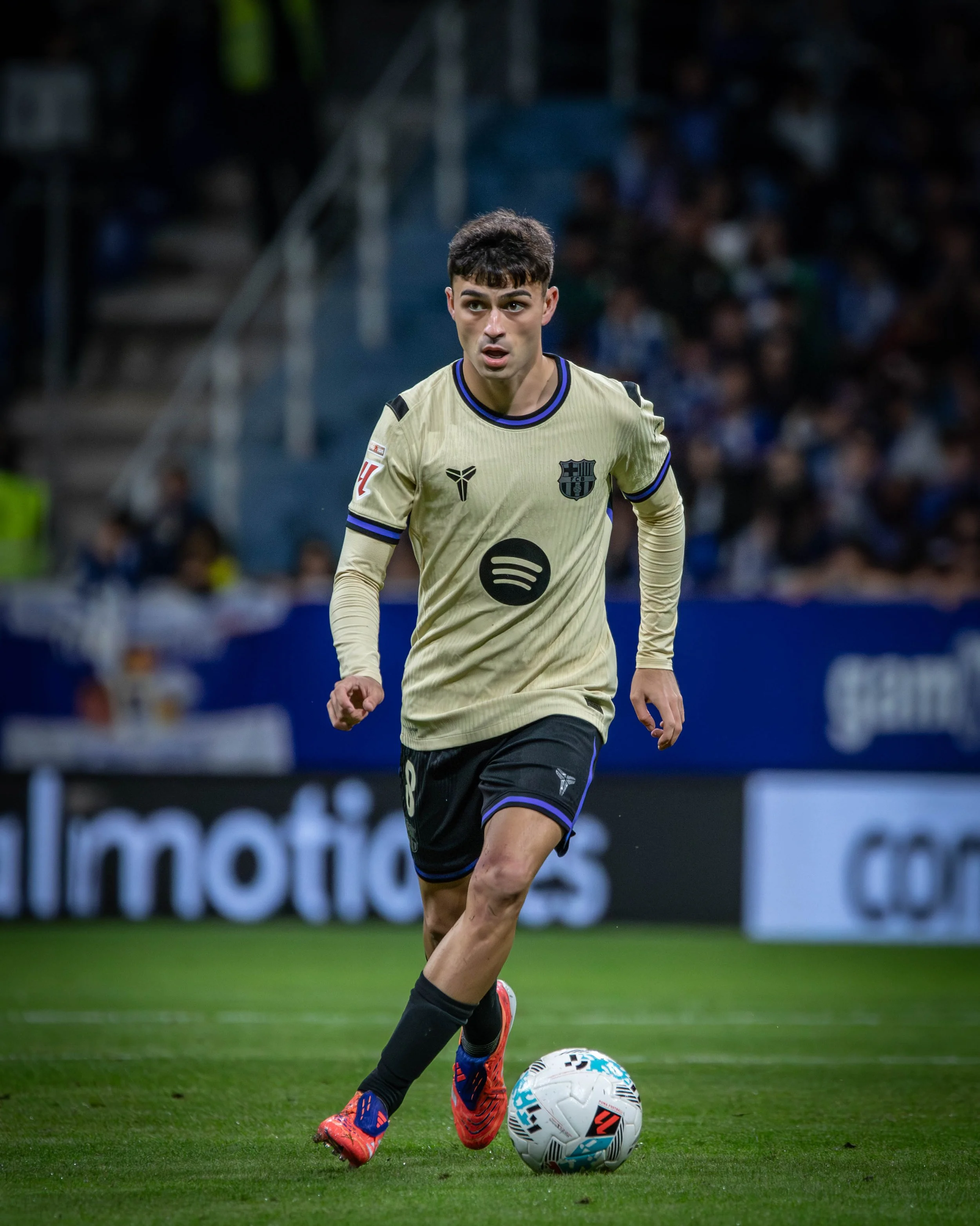 A soccer player in a beige jersey with black shorts is running on the field with a soccer ball, in a stadium filled with spectators.