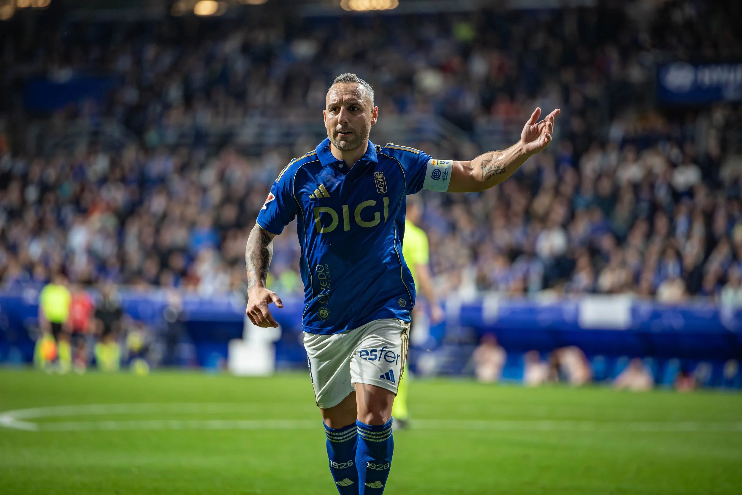 A soccer player in a blue uniform with white shorts and blue socks on a soccer field during a match, with a crowd in the stadium background.