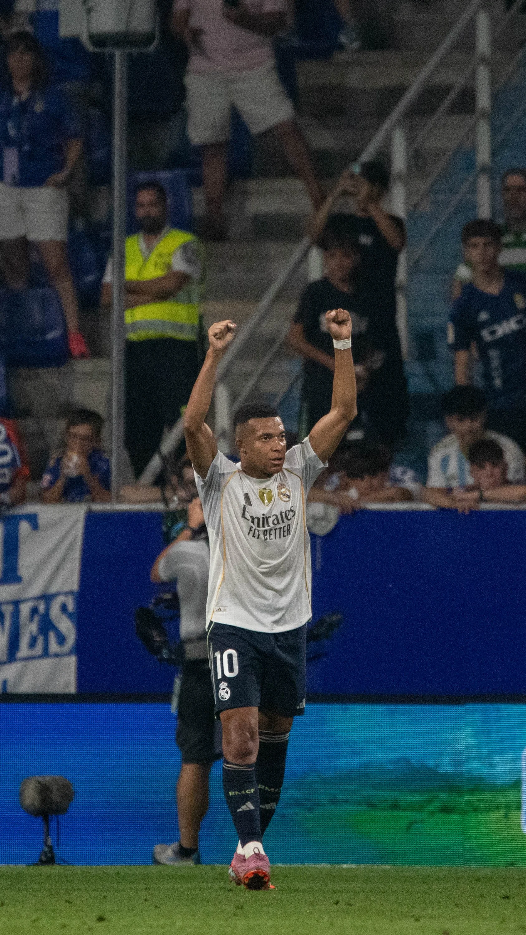 A soccer player celebrating on the field with arms raised, wearing a white jersey with the number 10 and a Real Madrid logo, during a match at night.