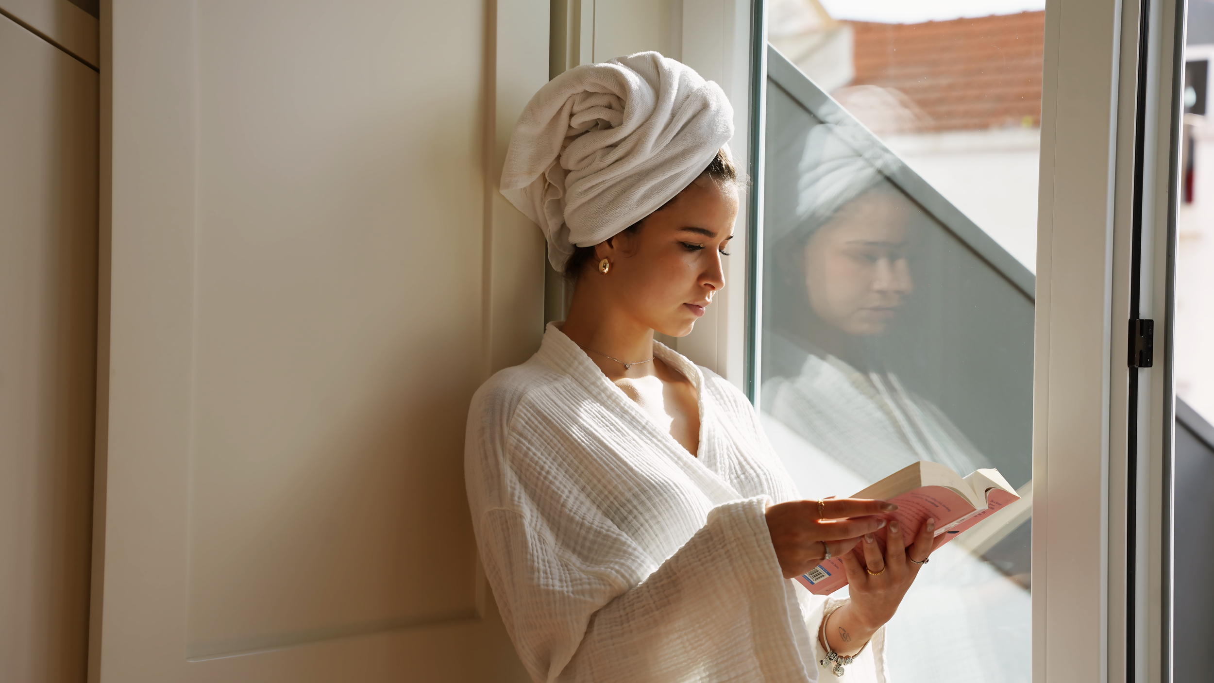 Cama com lençol branco, duas almofadas brancas, um cartão com a palavra "WELCOME" e uma flor azul ao lado, ao lado de uma parede com sombra de janela interferindo na iluminação