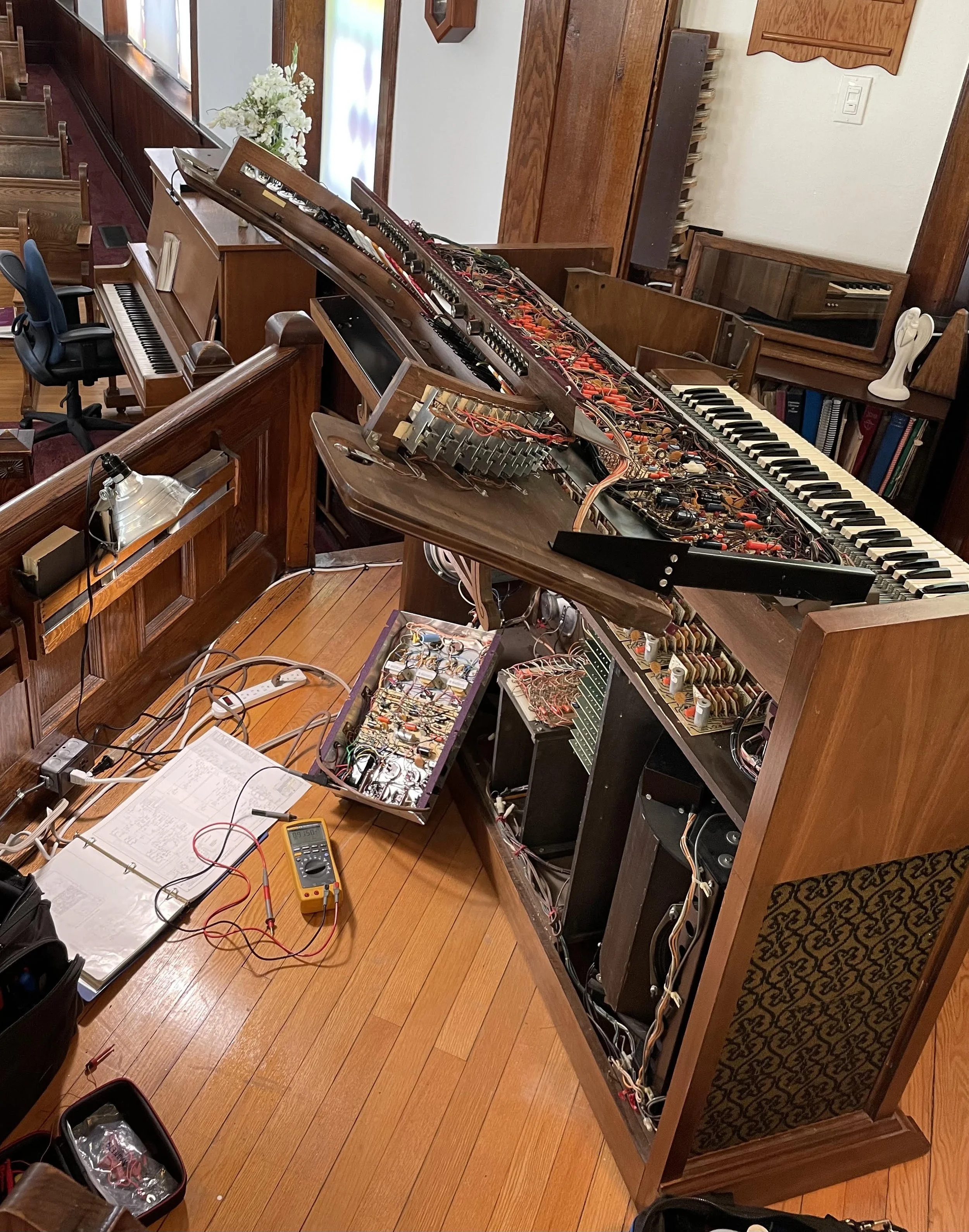 A wooden church podium with a disassembled vintage electronic organ on top of it, revealing internal wiring and components. Surrounding the organ are other musical instruments, books, and electronic equipment, indicating a repair or maintenance workspace inside a church or music room.