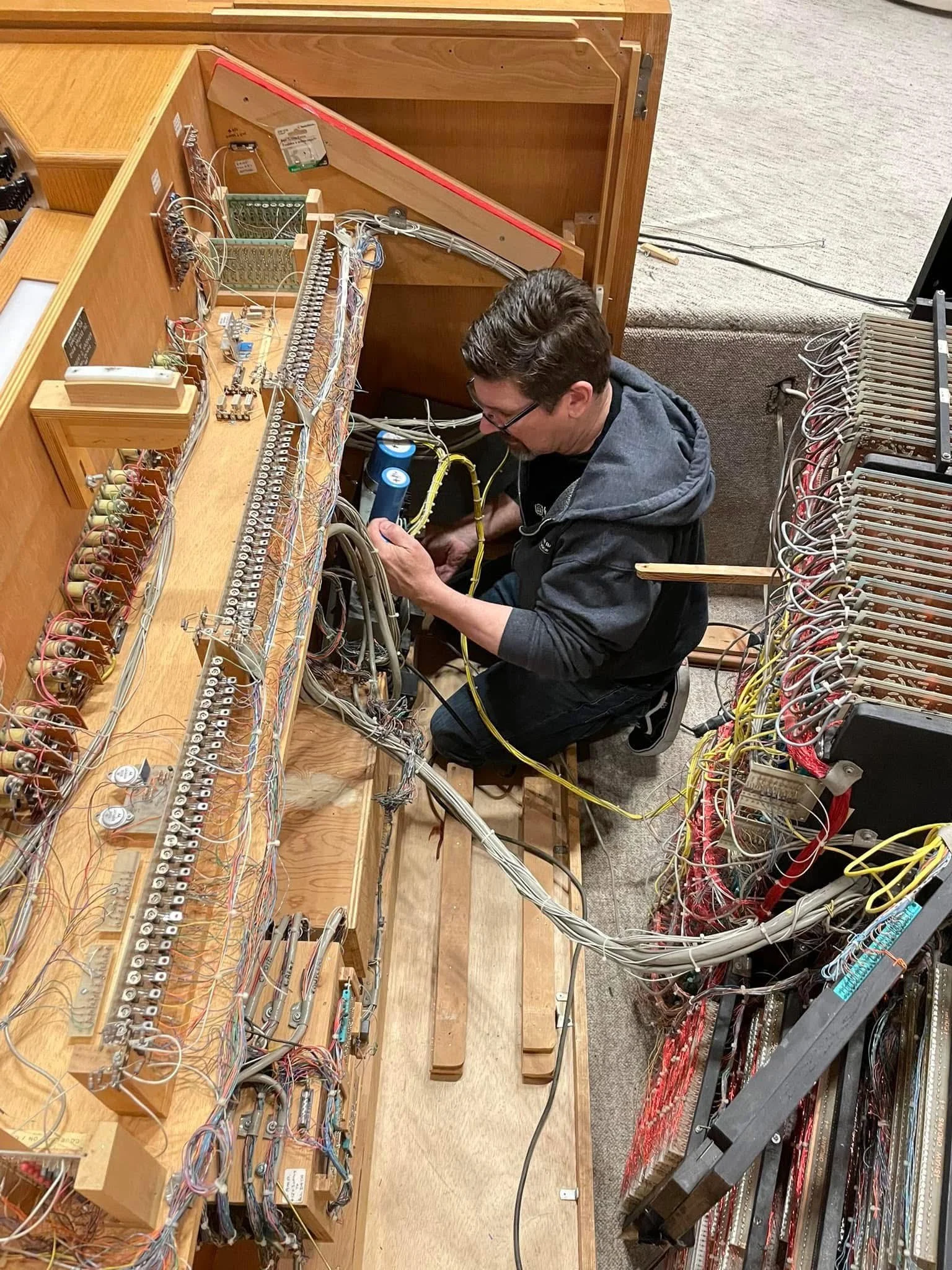 A man working on a large vintage mechanical organ with many wires and components, in a room with a carpeted floor.