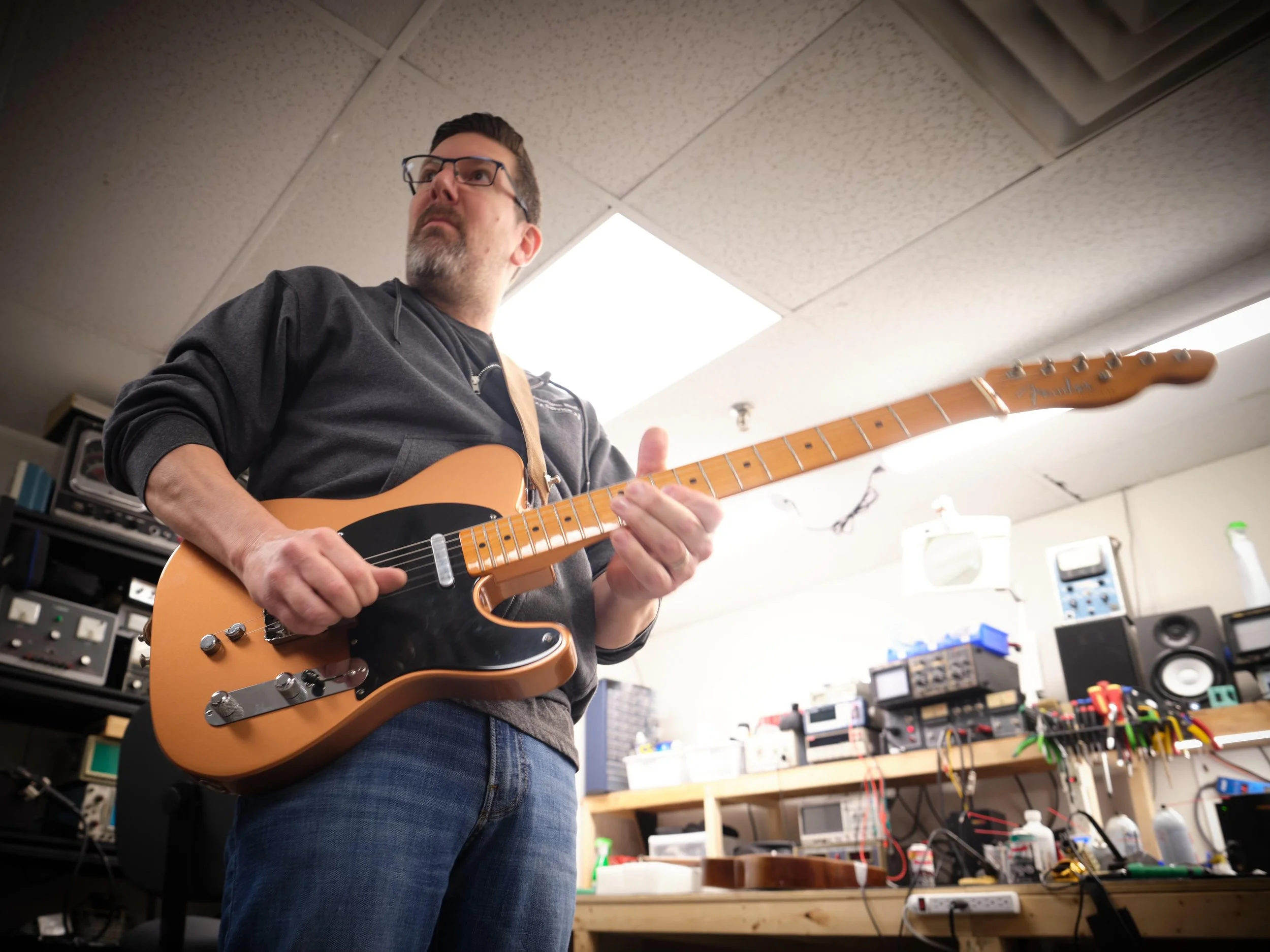 A man wearing glasses and a dark hoodie playing an orange electric guitar in a music or electronics workshop.