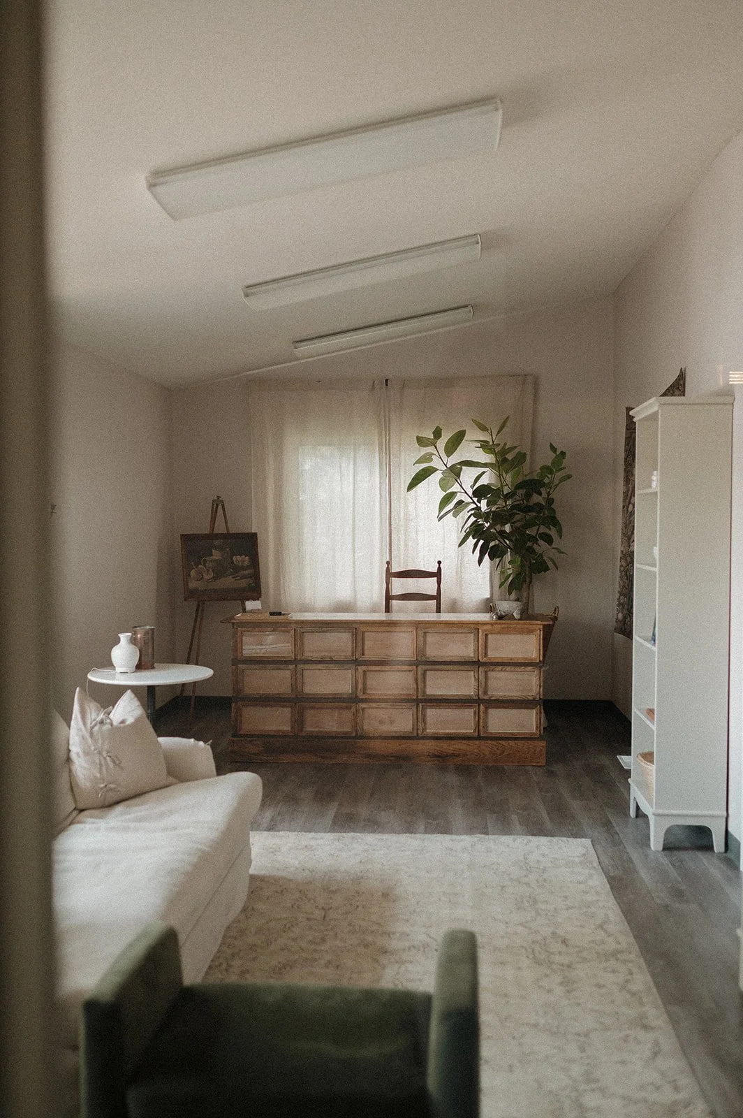 Yinergys Lobby, a room with a sofa, cushions, a window with curtains, a small table with vases, a chest of drawers, a chair, a large plant, and a painting on an easel.