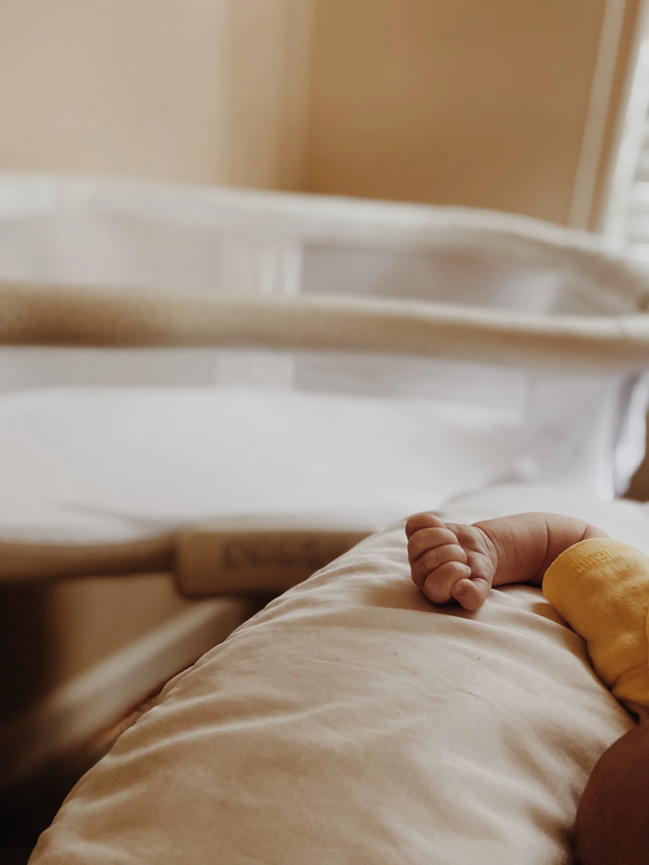 Close-up of a newborn baby's hand resting on a hospital bed, with a yellow hospital wristband visible, and a hospital bassinet in the background.