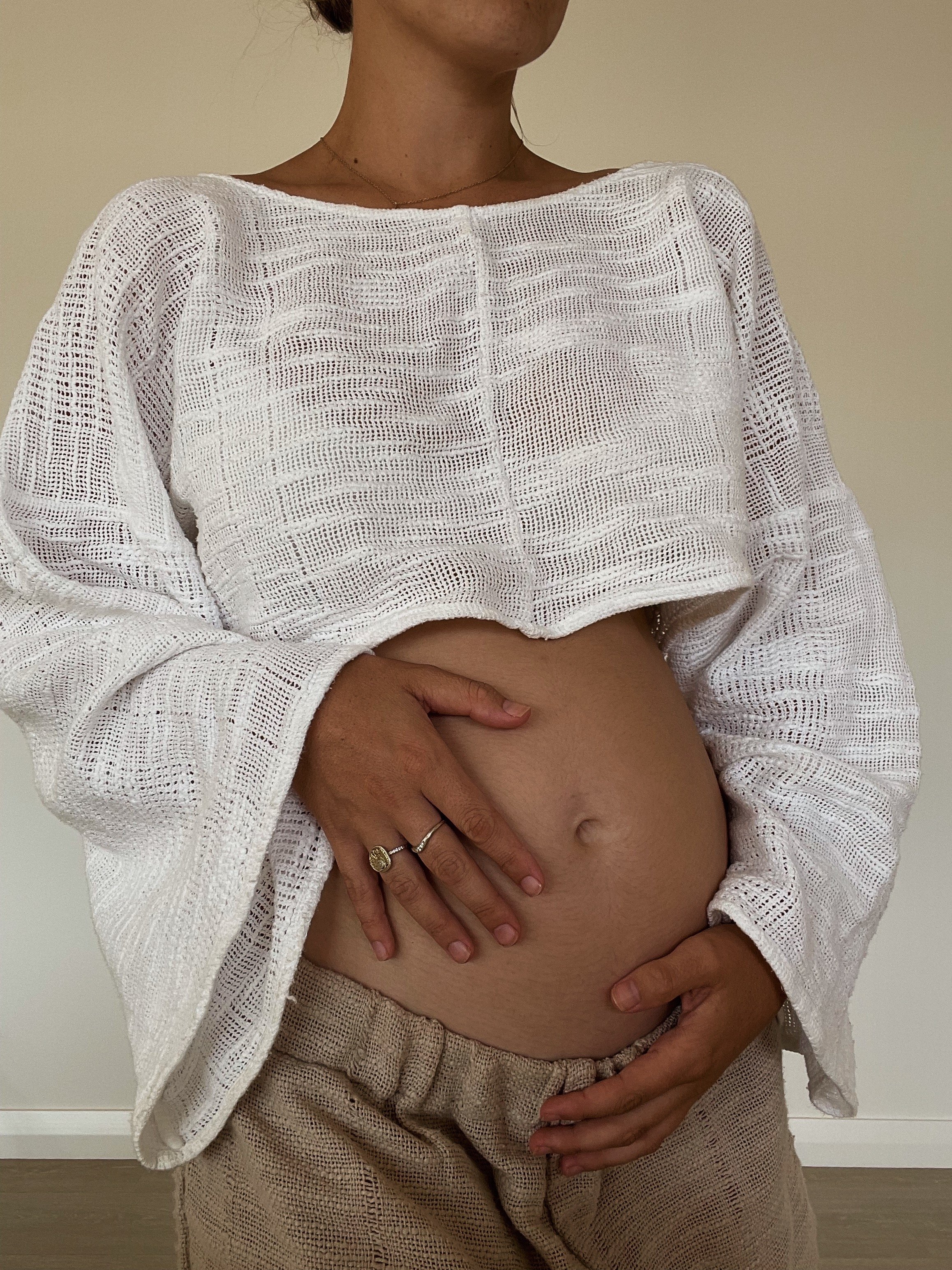 A woman wearing a white, loose-knit, long-sleeve top lifting it to reveal her pregnant belly, with her hands gently cradling her stomach.
